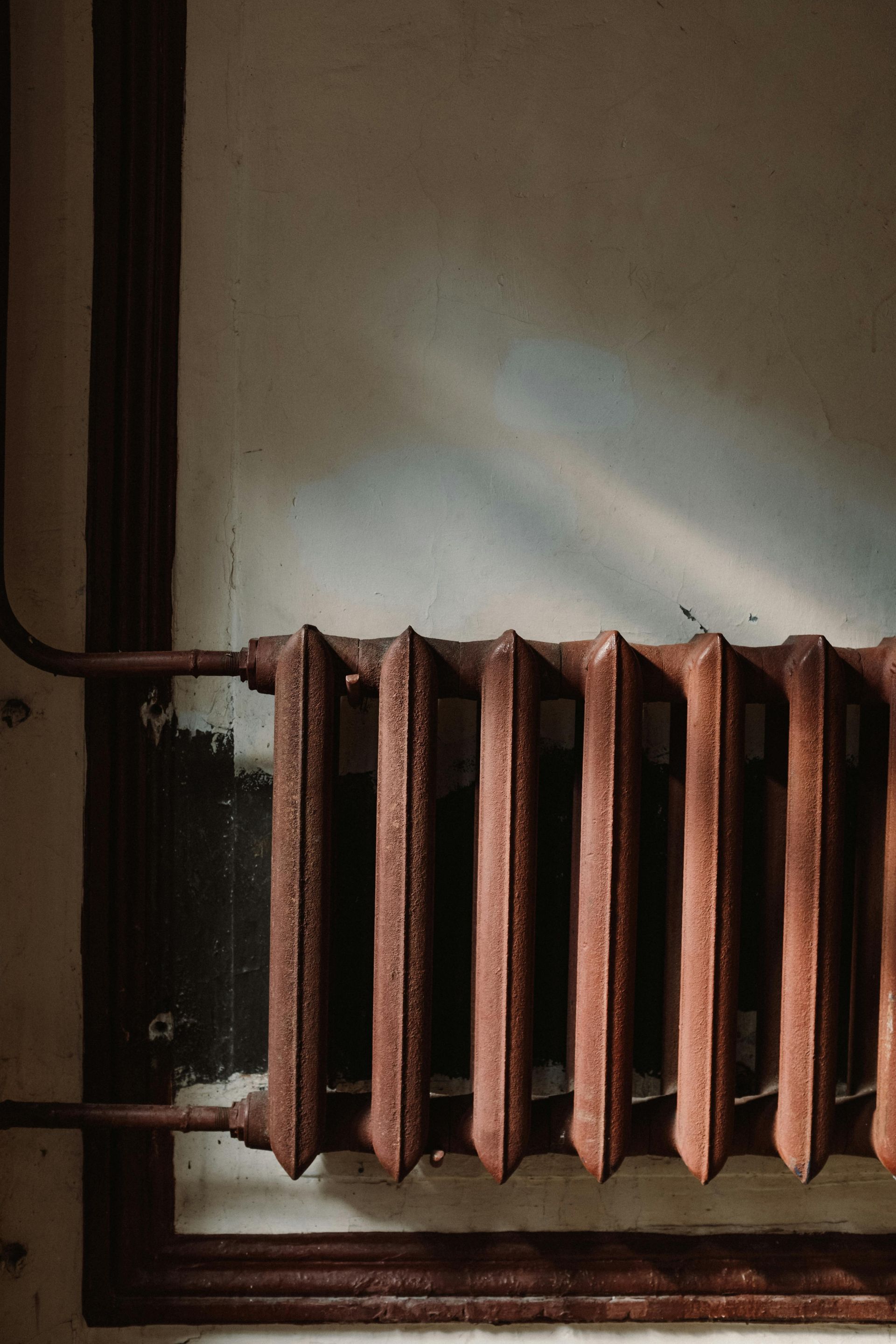 Rusty, vertical radiator against a light-streaked, aged wall. Dark frame and pipes are visible.