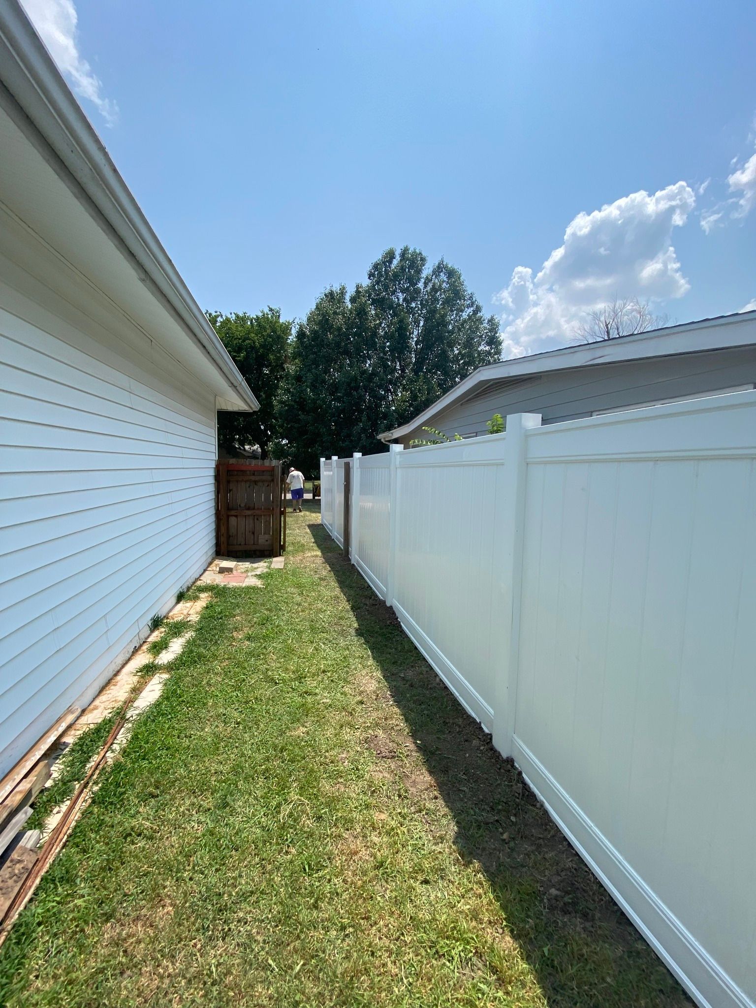 Narrow grassy space between white house and fence under blue sky.