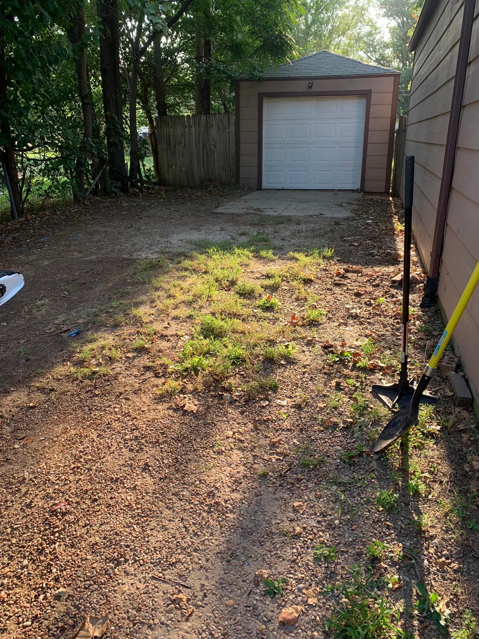 Dirt driveway leading to a garage with a closed white door, flanked by a fence and a house.