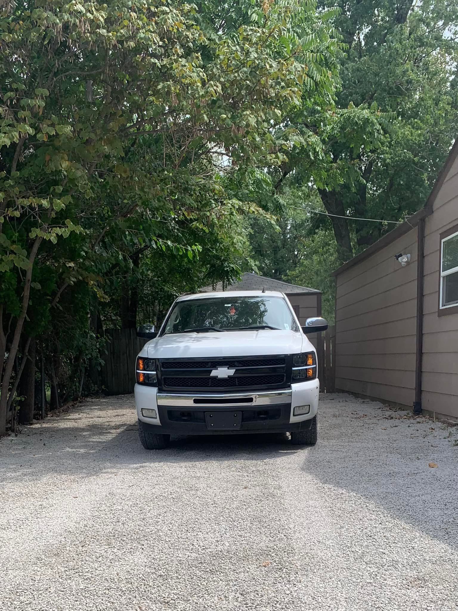 White pickup truck parked on gravel driveway, surrounded by trees and a brown building.