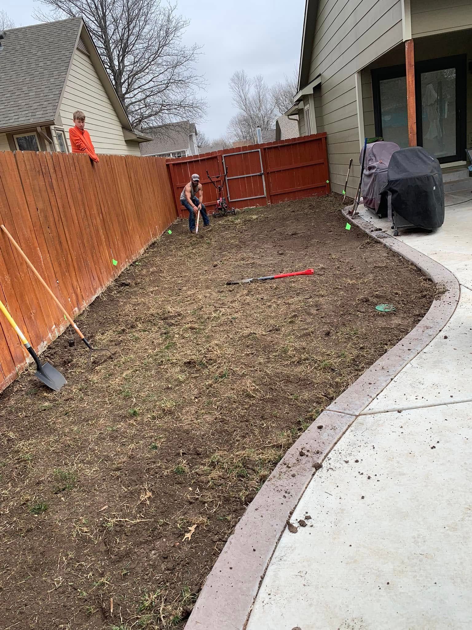 Workers in a backyard prepping garden bed. Brown fence, concrete edging, lawn covered in mulch.