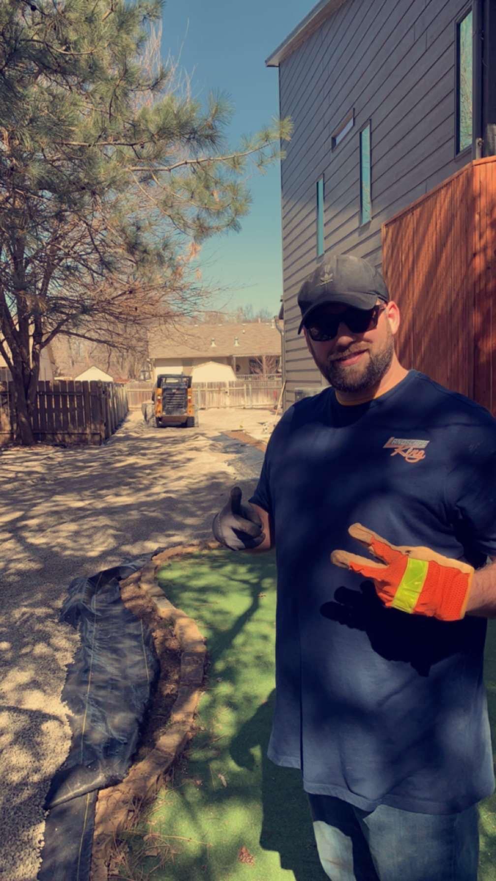 Man in sunglasses, holding up two fingers, stands outside near construction site with a small digger on a sunny day.