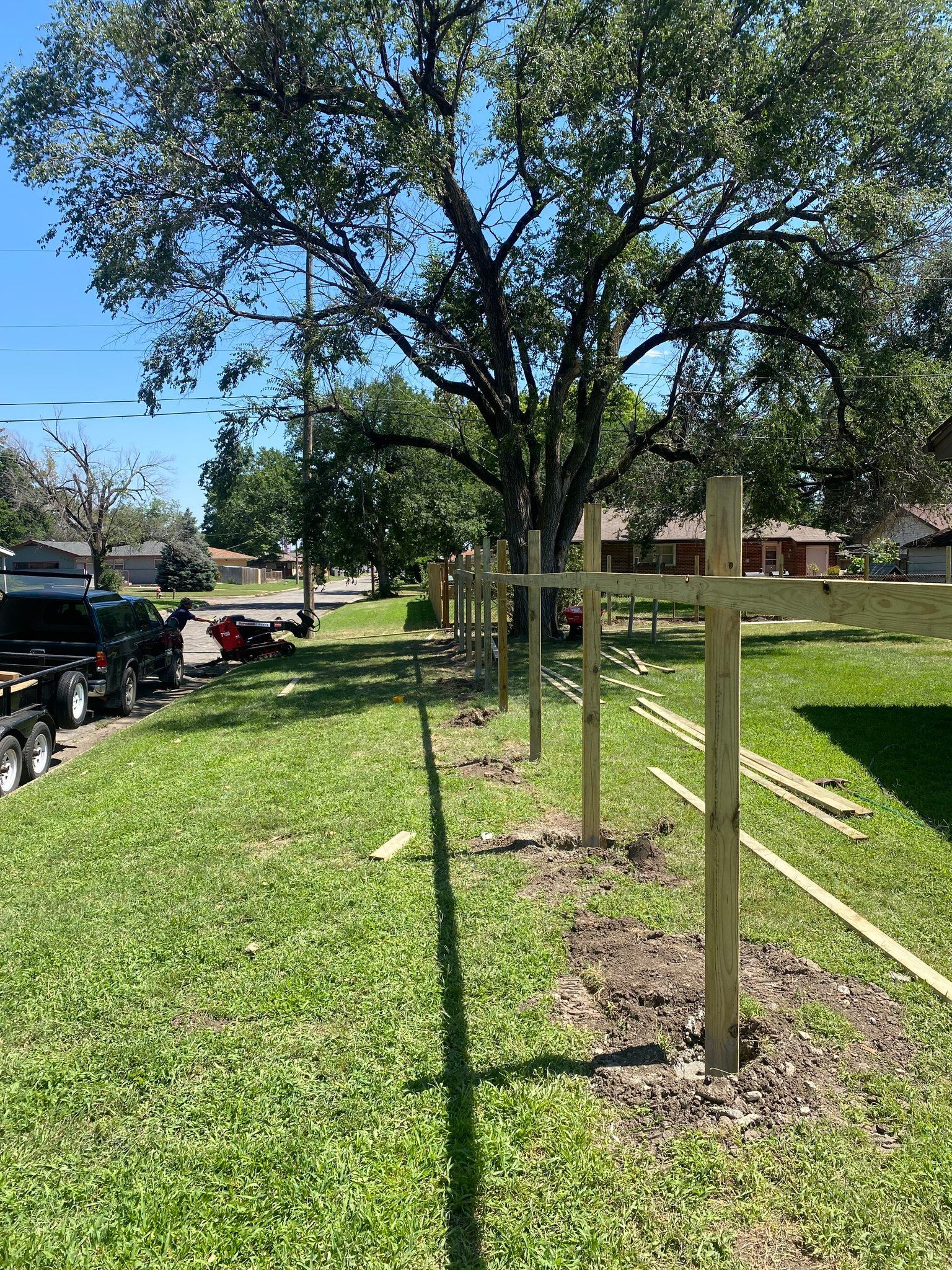 Wooden fence posts installed along a grassy lawn, tree in background, sunny day.
