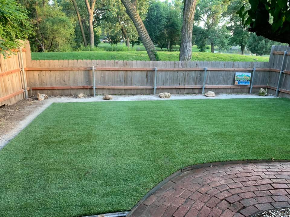 Green artificial lawn in a backyard with a wooden fence, stones, and a brick patio.