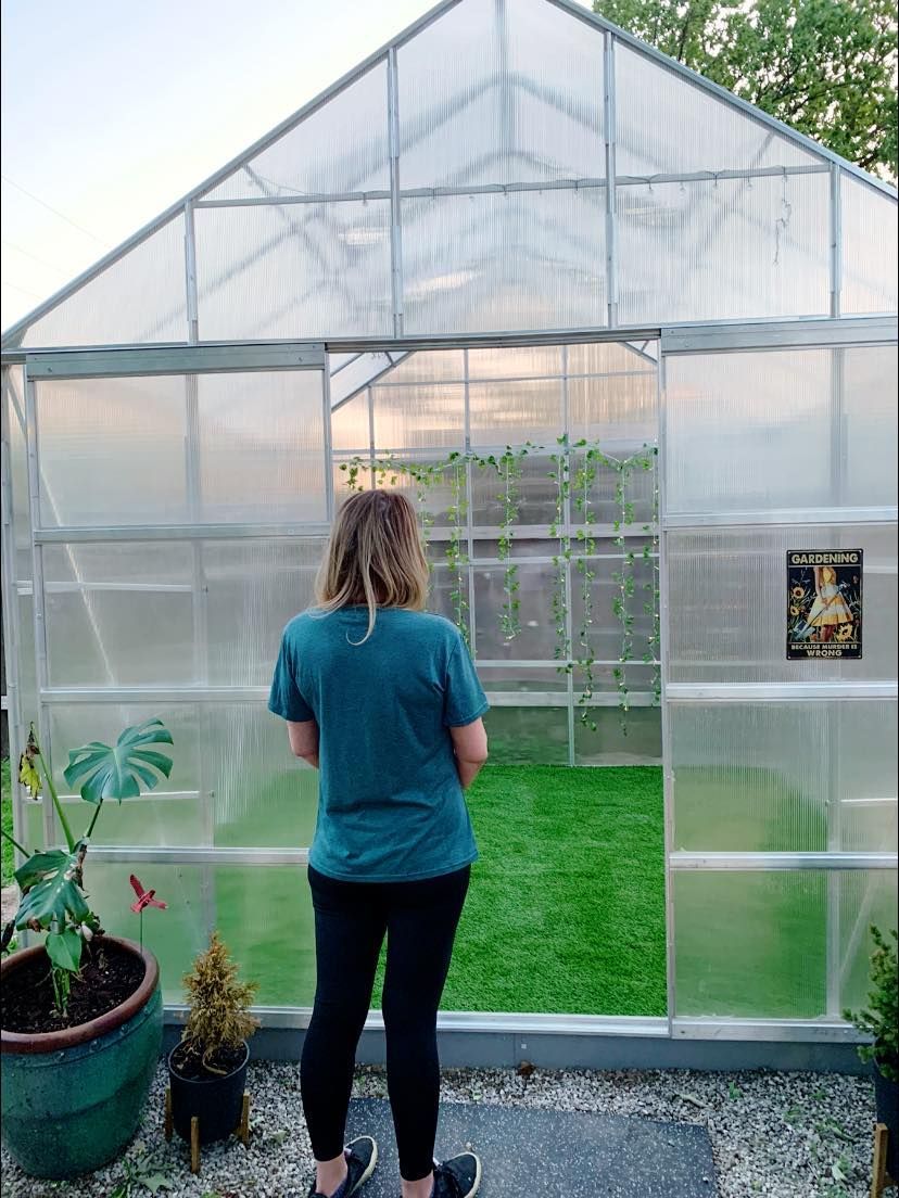 Woman stands in front of greenhouse, looking inside; green lawn visible.