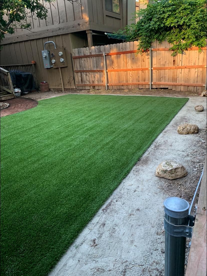 Backyard with green turf and a wood fence. A concrete path borders the turf, with some decorative rocks.