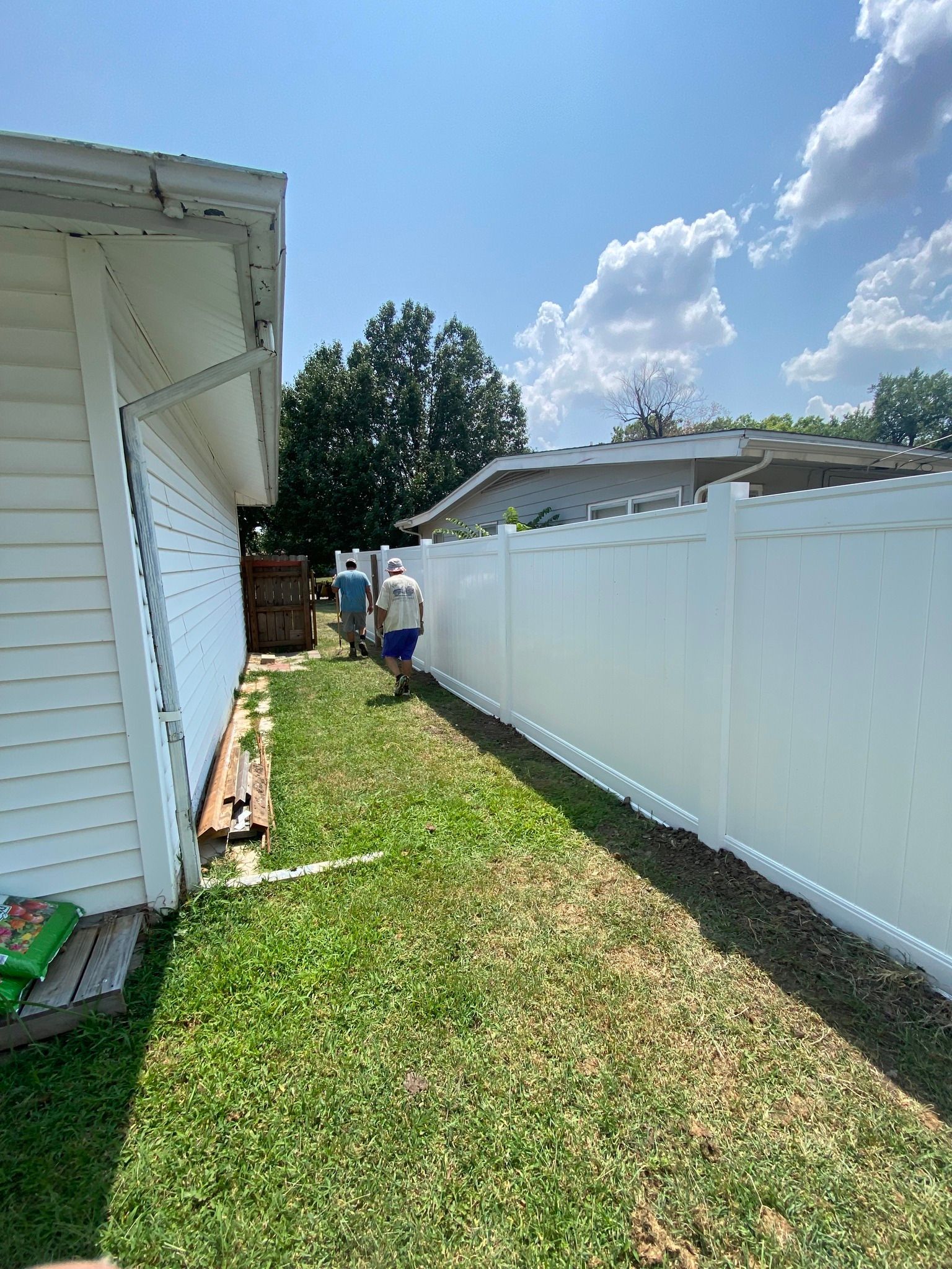 Two men walking between a white fence and a white building, grassy area, blue sky.