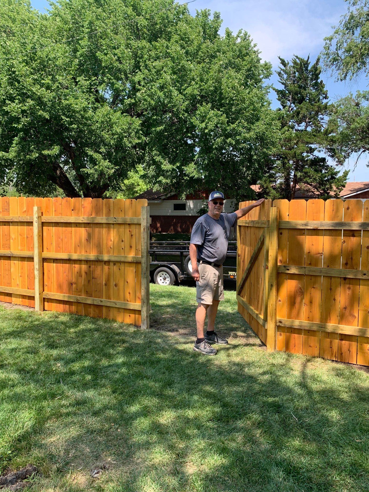 Man standing in an open gate of a new wood fence, green grass and trees in the background.