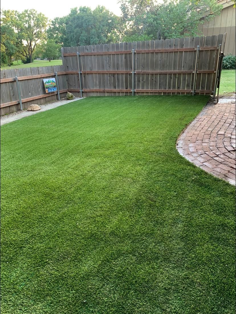 Lush green artificial turf backyard with a wooden fence and brick patio.