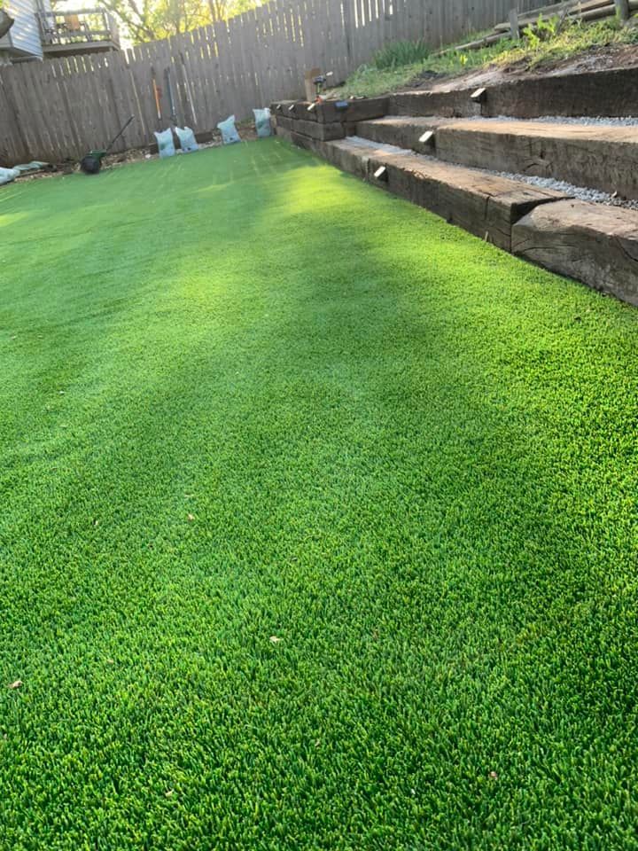 Lush green artificial lawn with wooden steps leading uphill against a wooden fence, bathed in sunlight.