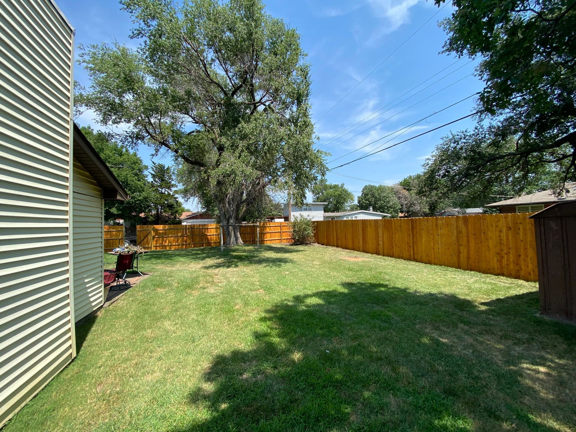 Backyard with a green lawn, brown wooden fence, and large tree under a blue sky.