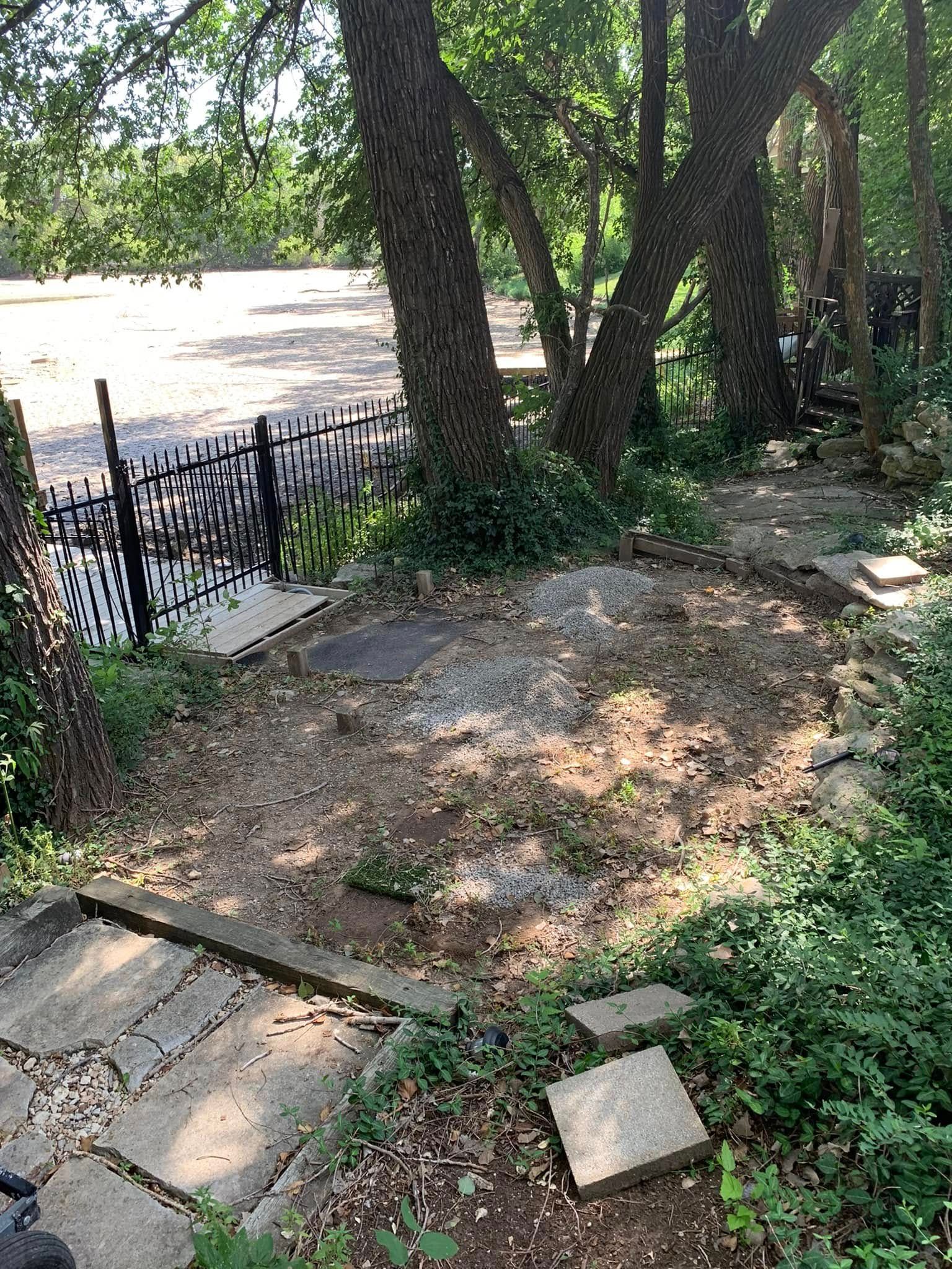 A small clearing with trees, stone steps, and a wrought-iron fence bordering a river.