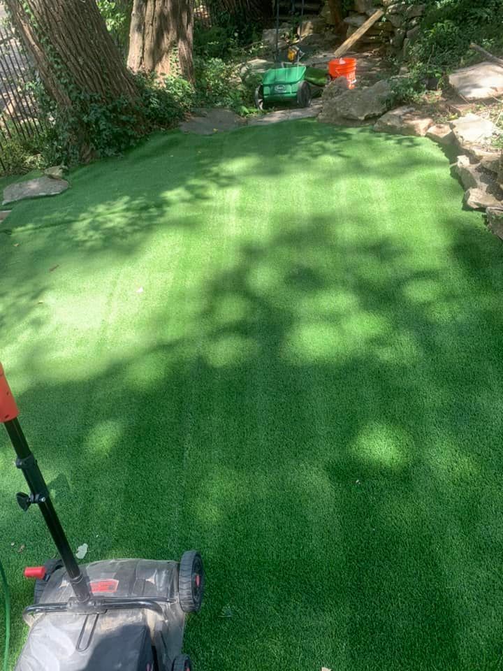 A lawnmower on artificial grass in a backyard setting. The grass is bright green, and the scene is sunny.