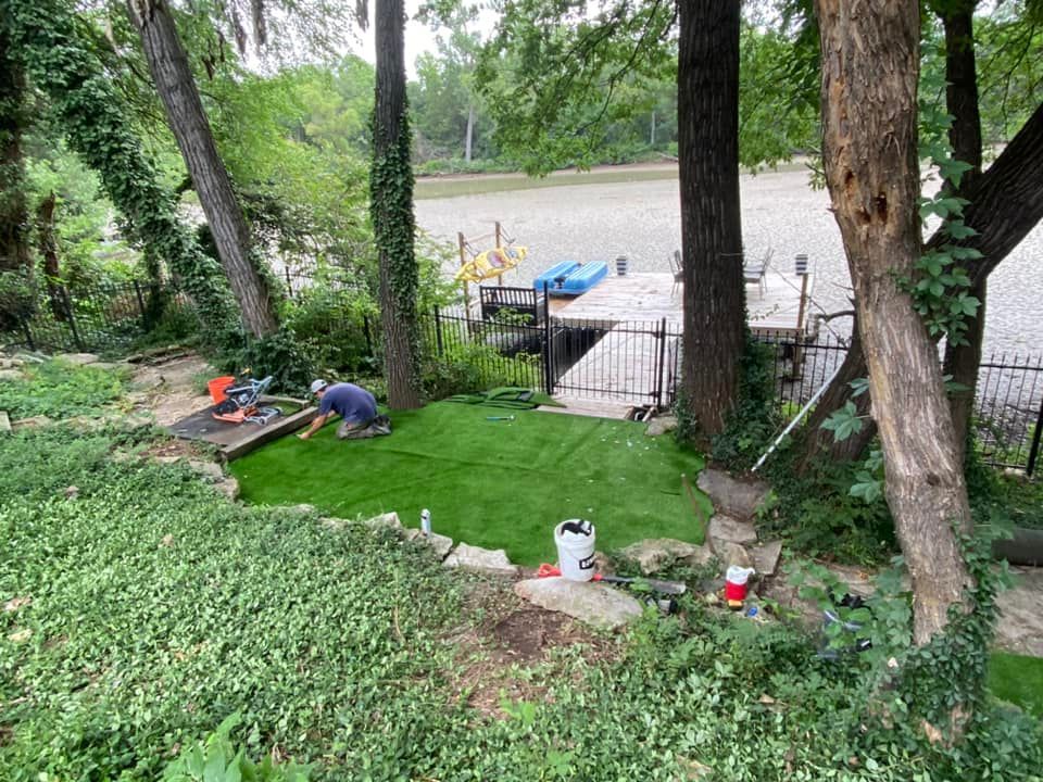 Man installing artificial grass by a river, surrounded by trees and greenery.