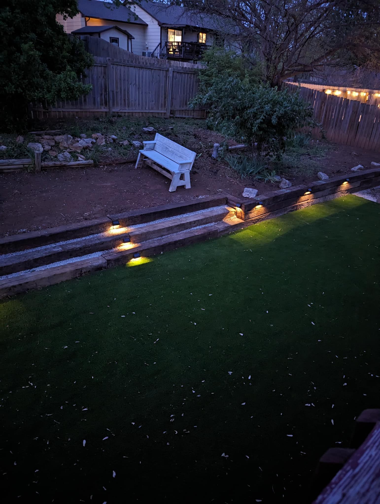 Backyard at dusk: green lawn, stone steps with lights, white bench, string lights, house in the background.