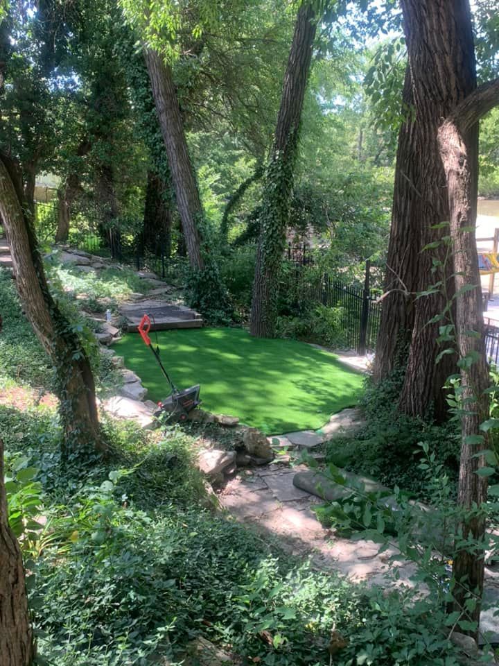 Pathway leading to small green artificial lawn surrounded by trees and foliage.