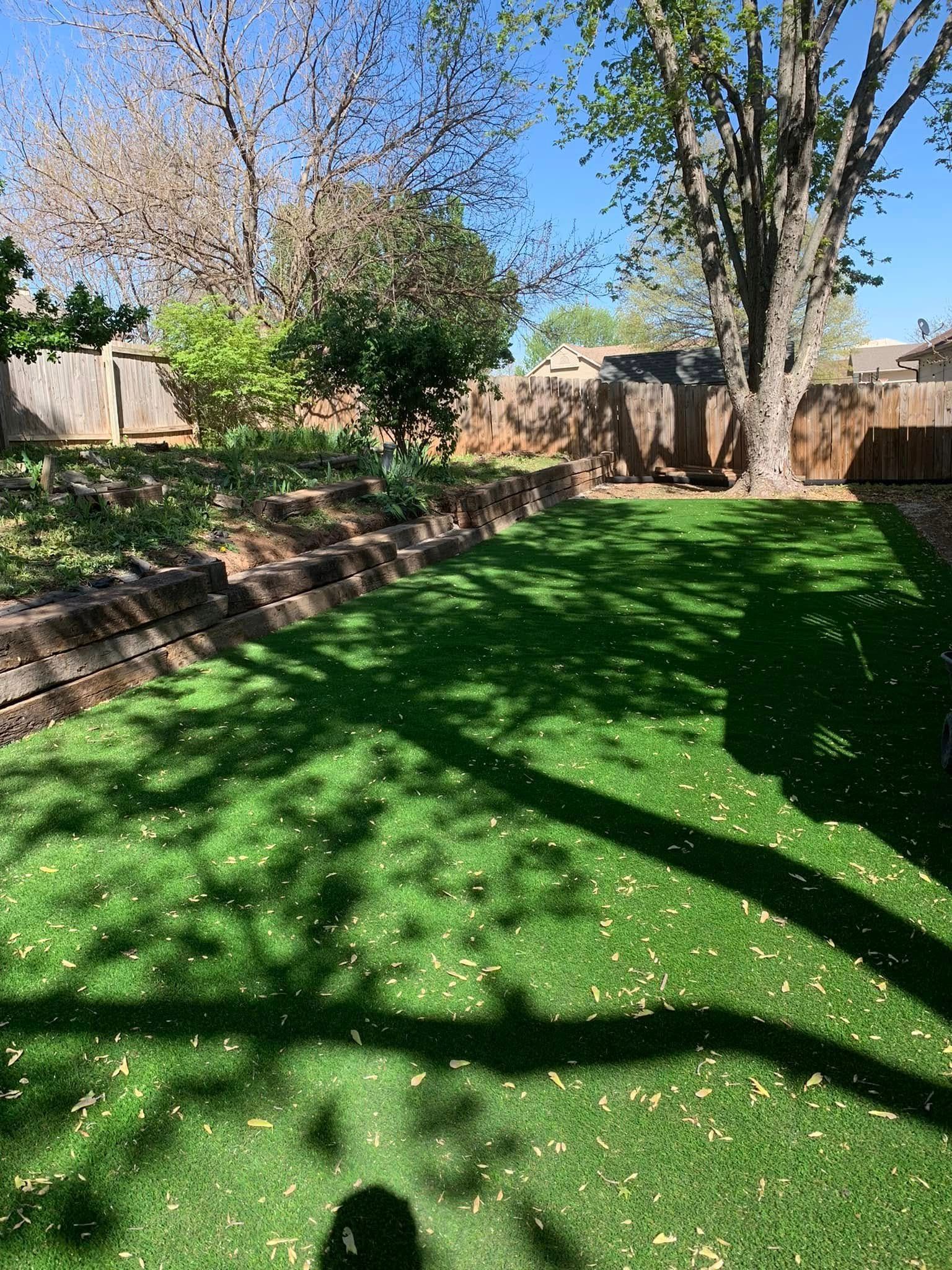 Green lawn in backyard with tiered wooden retaining wall, fence, and trees.