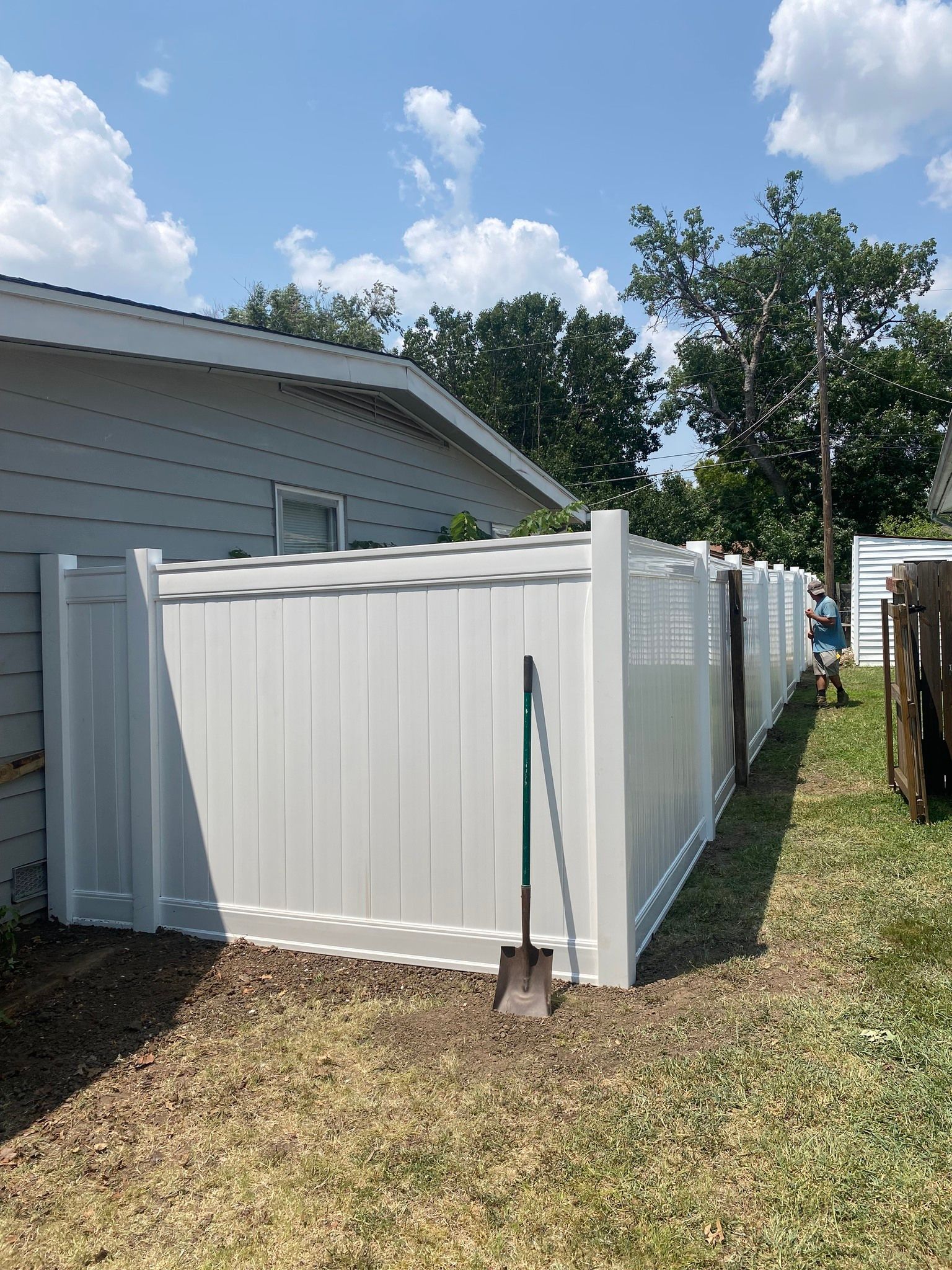 White vinyl fence alongside a building, shovel leans against it, sunny day.