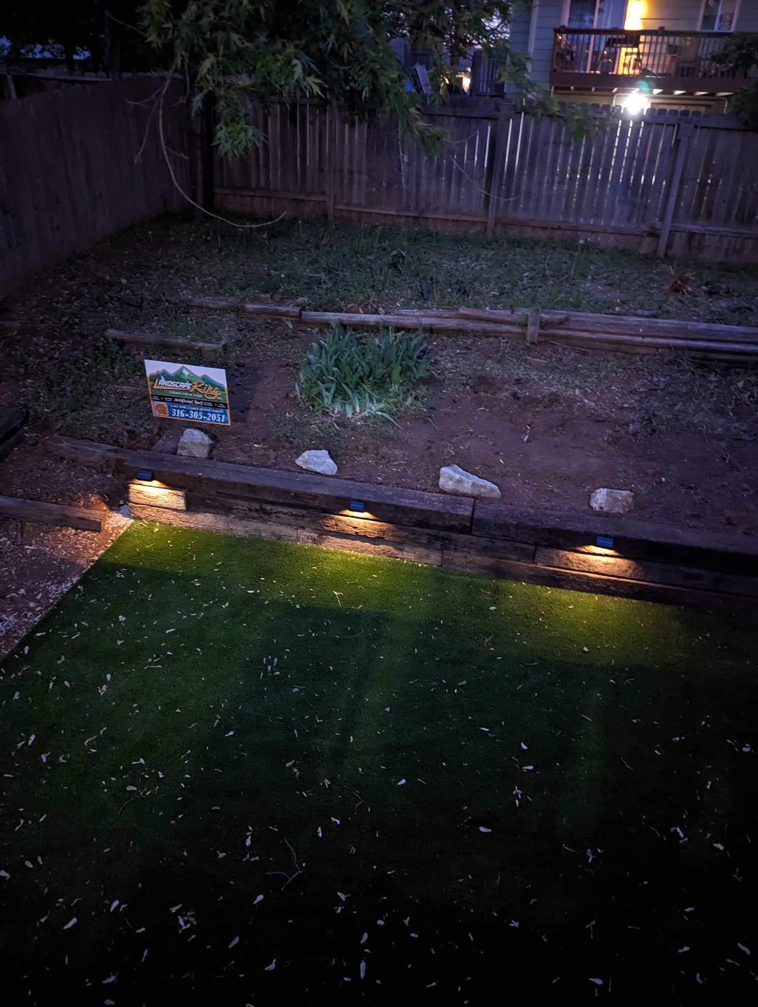 Backyard at night with landscape lighting illuminating the grass and a garden bed. Wooden fence and a house in the background.