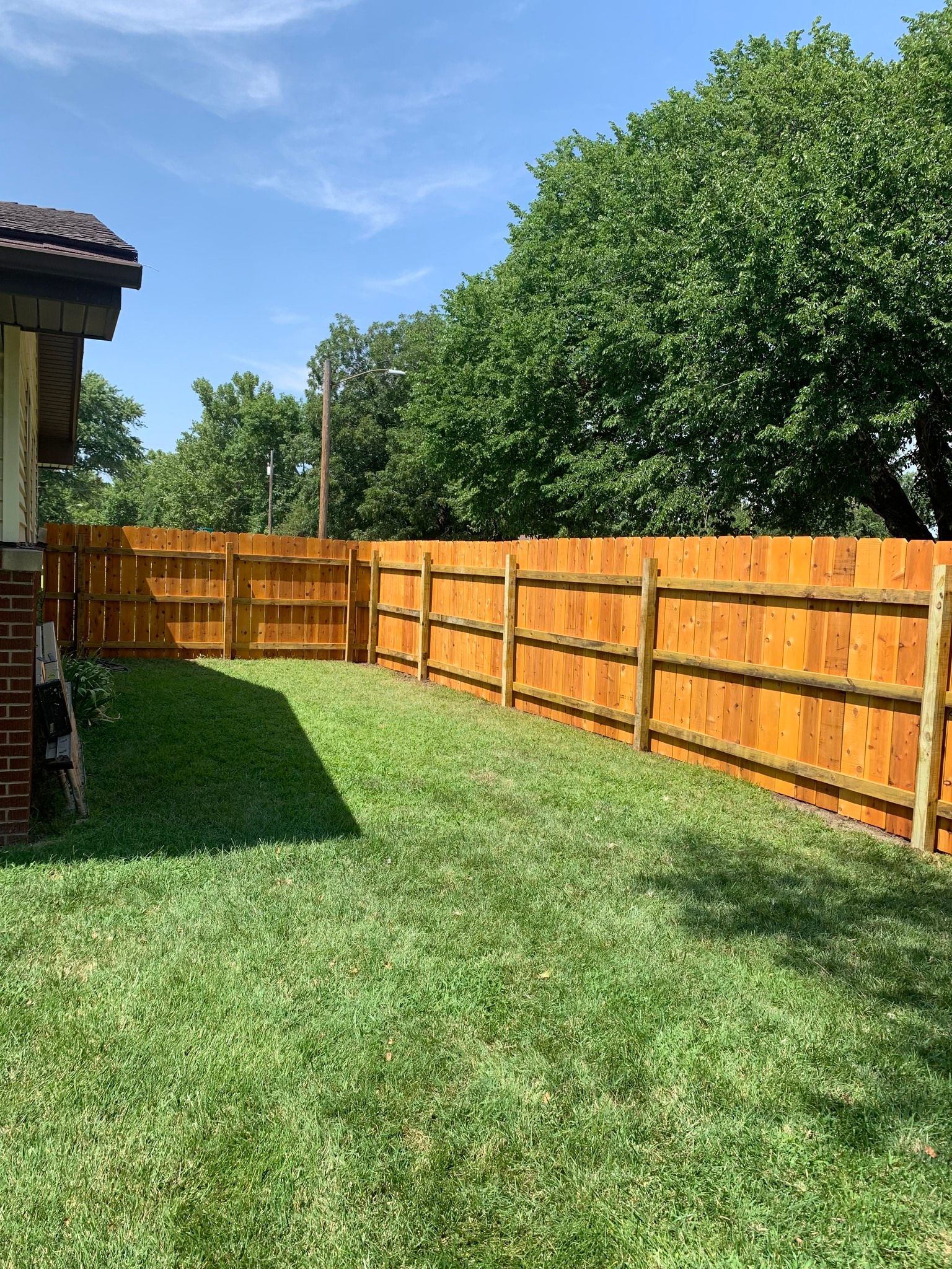 Wooden fence encloses a grassy yard, extending alongside a house with a brick facade, under a blue sky.