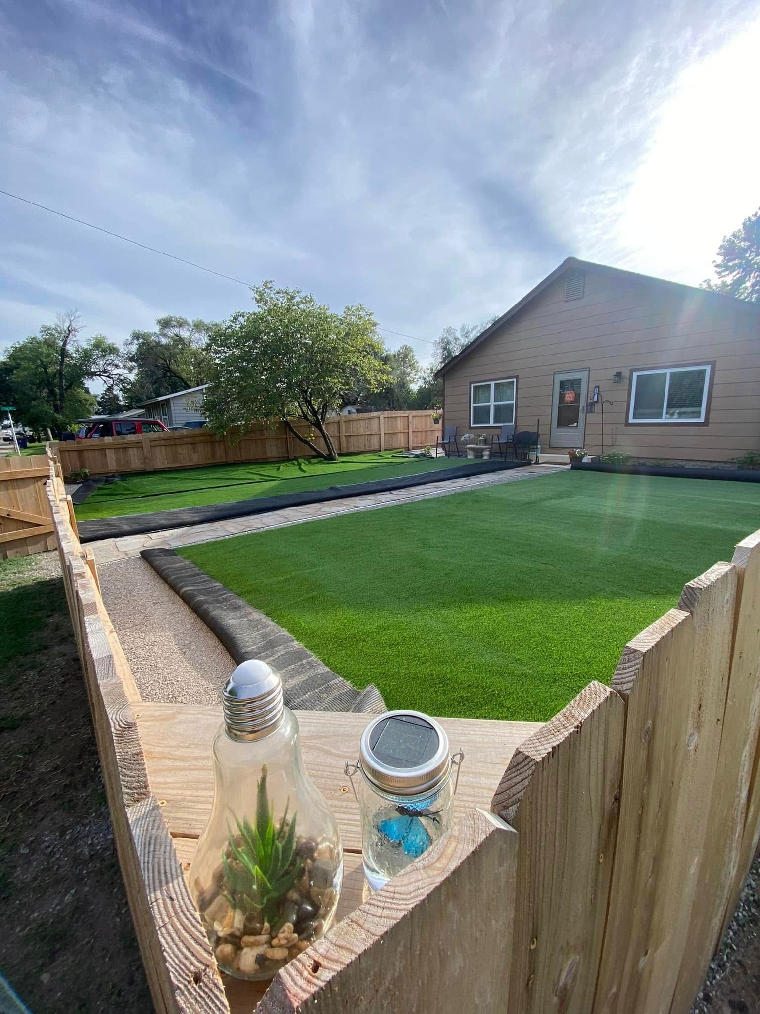Backyard with green lawn, tan fence, and a light-colored brick house under a partly cloudy sky.