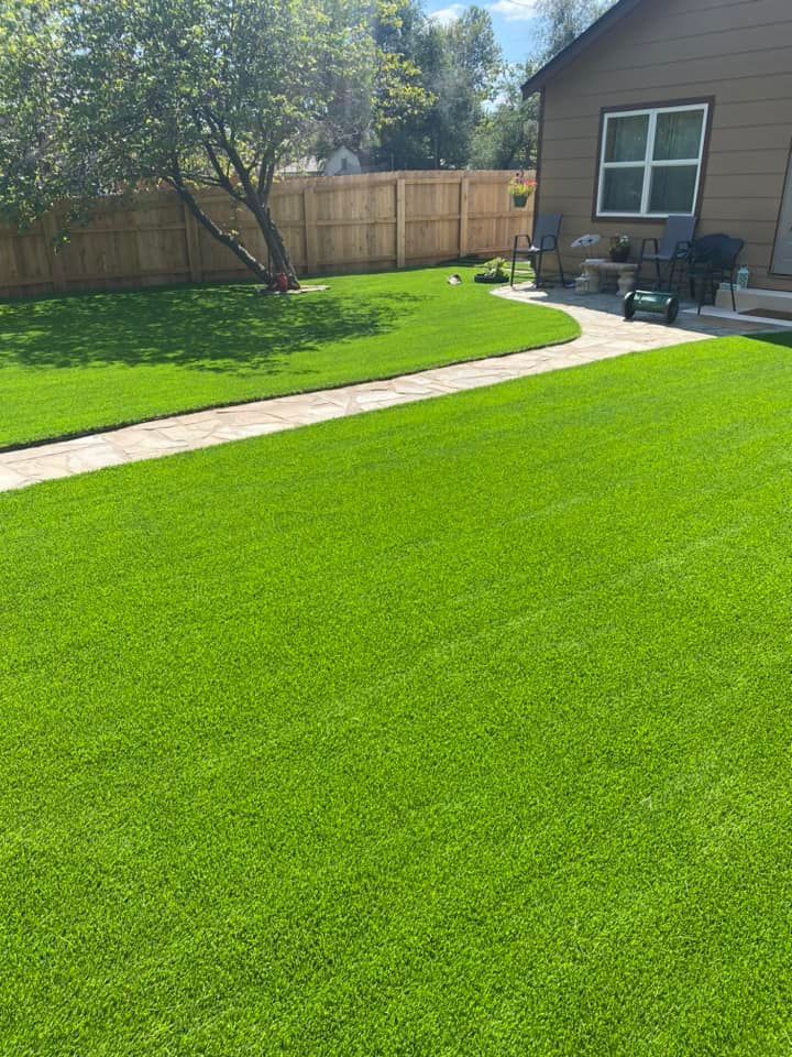 Lush green lawn with a stone path leading to a house. Wooden fence and tree in the background.