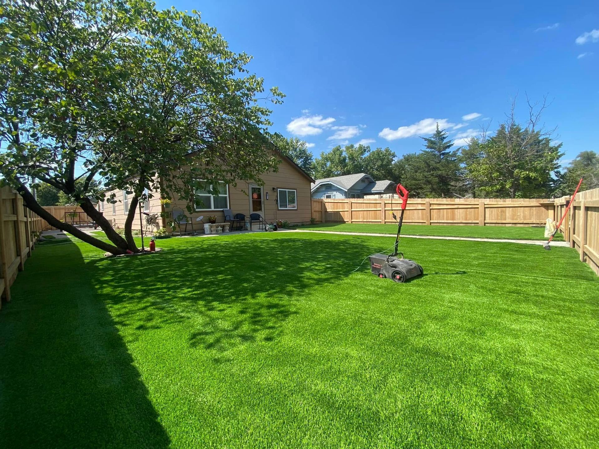Lush green lawn in a fenced backyard with a house and a tree, on a sunny day. A robotic mower is in the grass.
