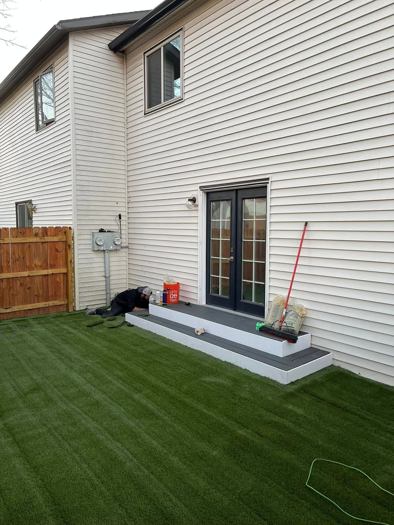 Back of a two-story house with a lawn. Steps lead to black French doors. White siding, wooden fence, and a red broom are visible.