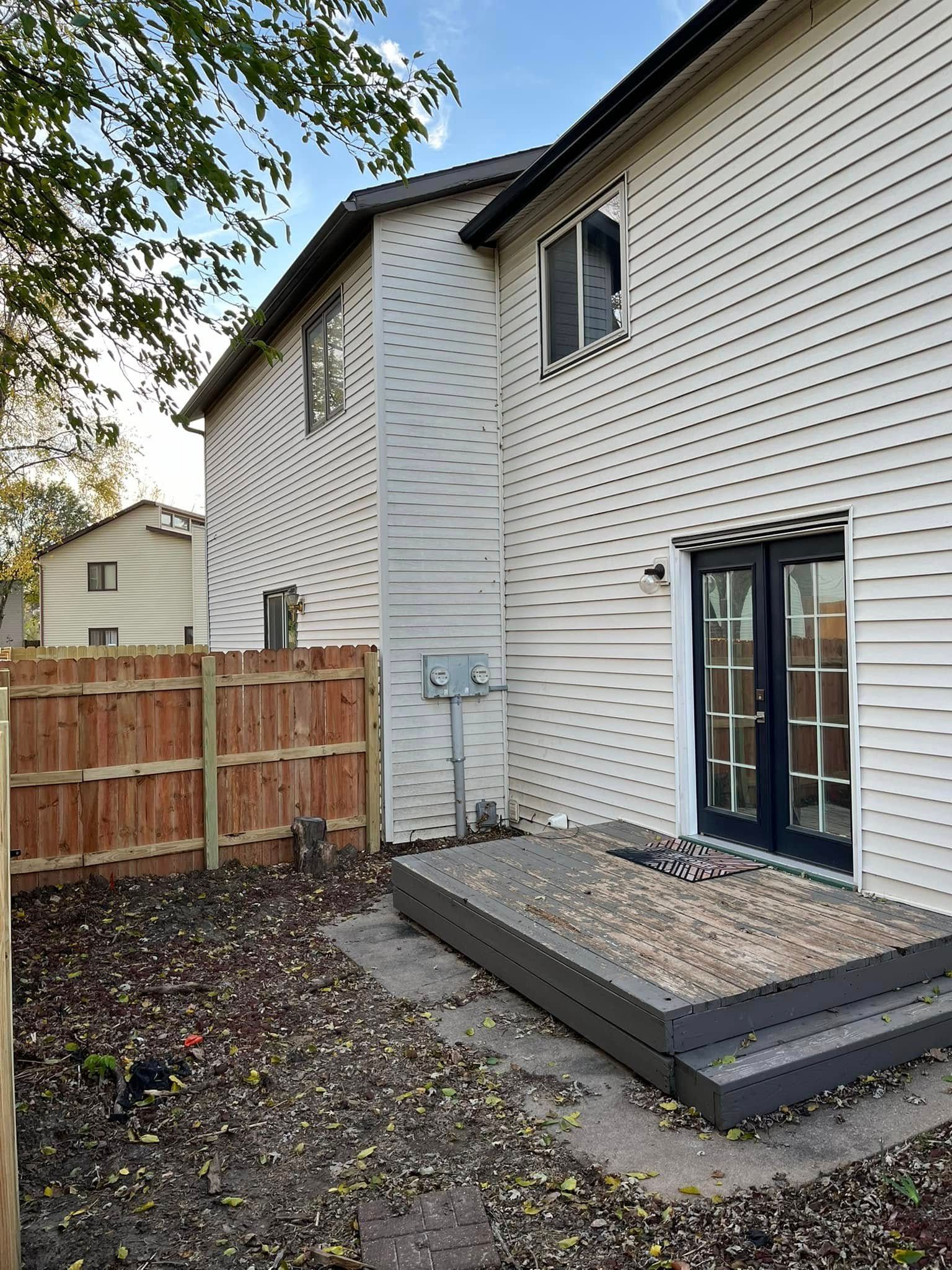 Back of a white house with a dark door, wooden deck, and a brown fence.