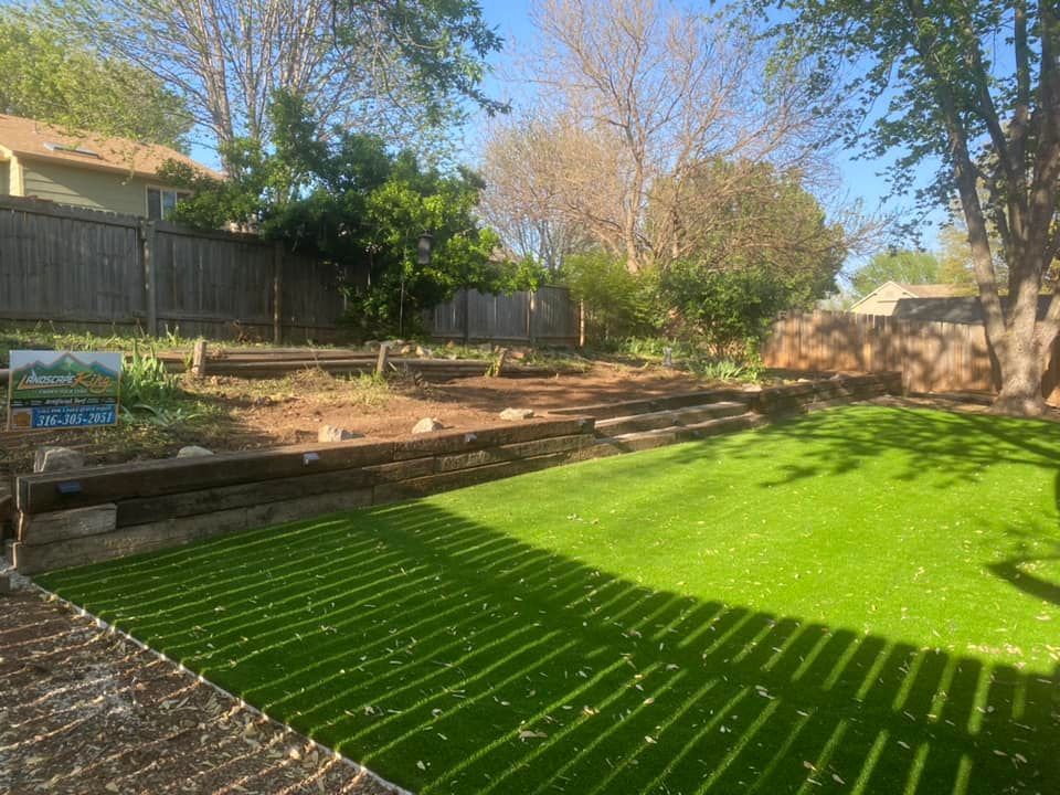 Green artificial turf lawn with wooden steps and retaining wall. Fenced backyard, sunny day.