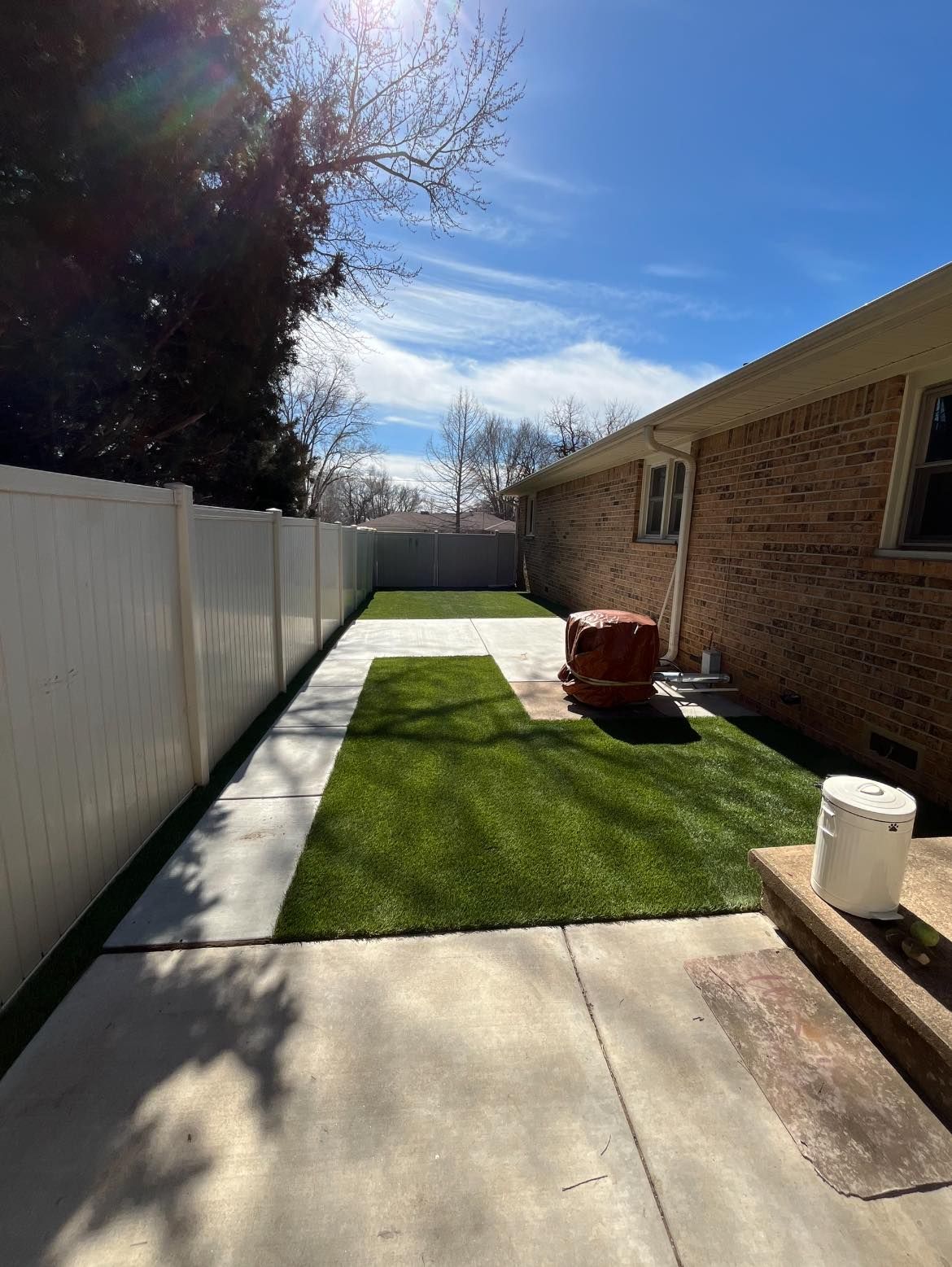Backyard with concrete patio, artificial turf, white fence, and brick house under a sunny sky.