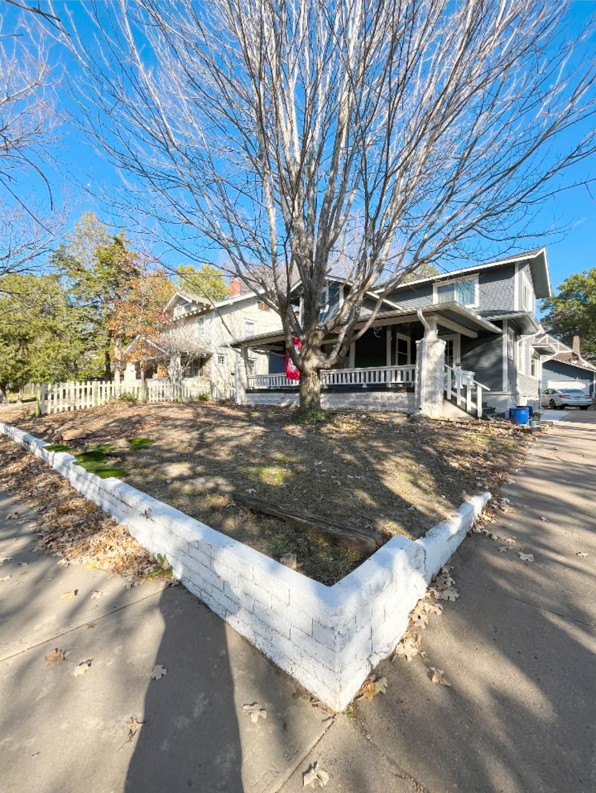 Two-story house behind a white brick wall with a large tree in front. Gray exterior, blue sky.