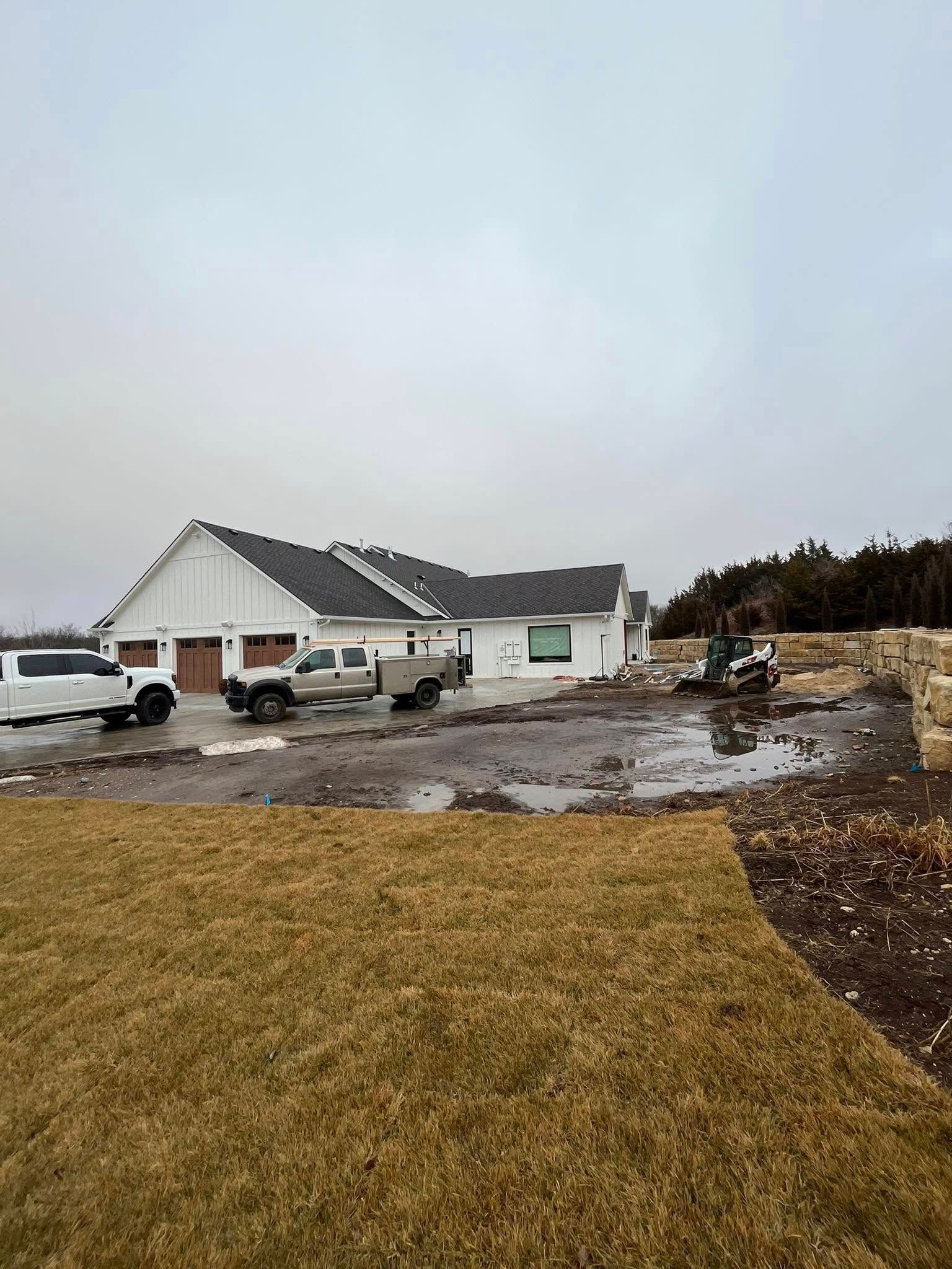 White house under construction, parked trucks, muddy yard, overcast sky.
