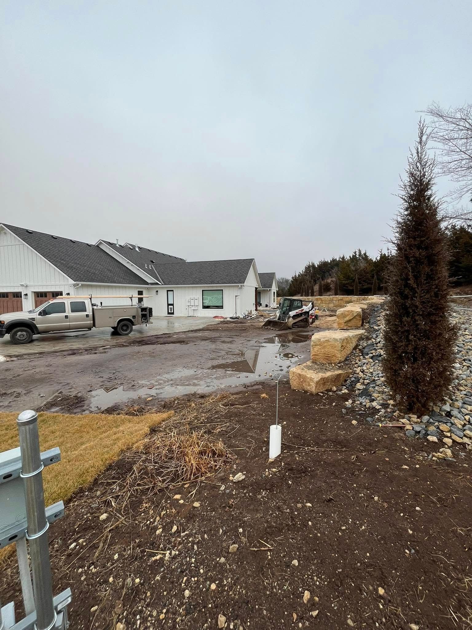 Muddy construction site with new white houses, truck, and retaining wall. Overcast sky.