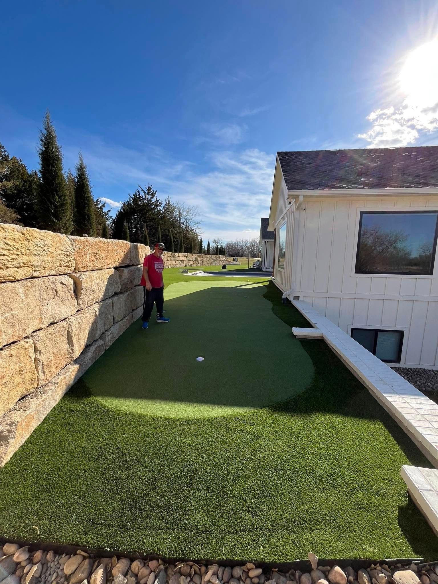 Man putting on a backyard green next to a stone wall and a white house on a sunny day.