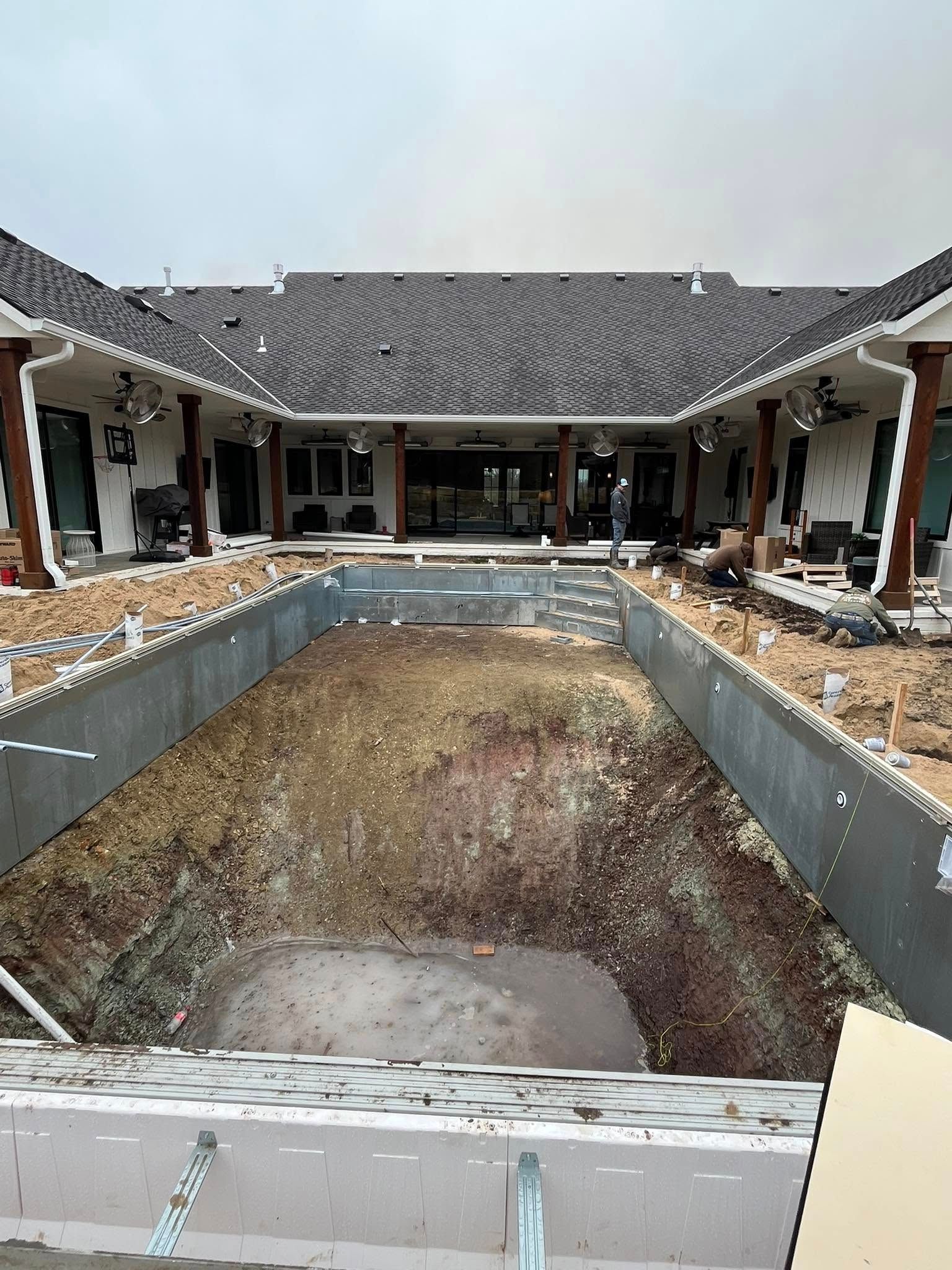 Empty pool under construction in front of a house with brown columns, white walls, and a dark roof.