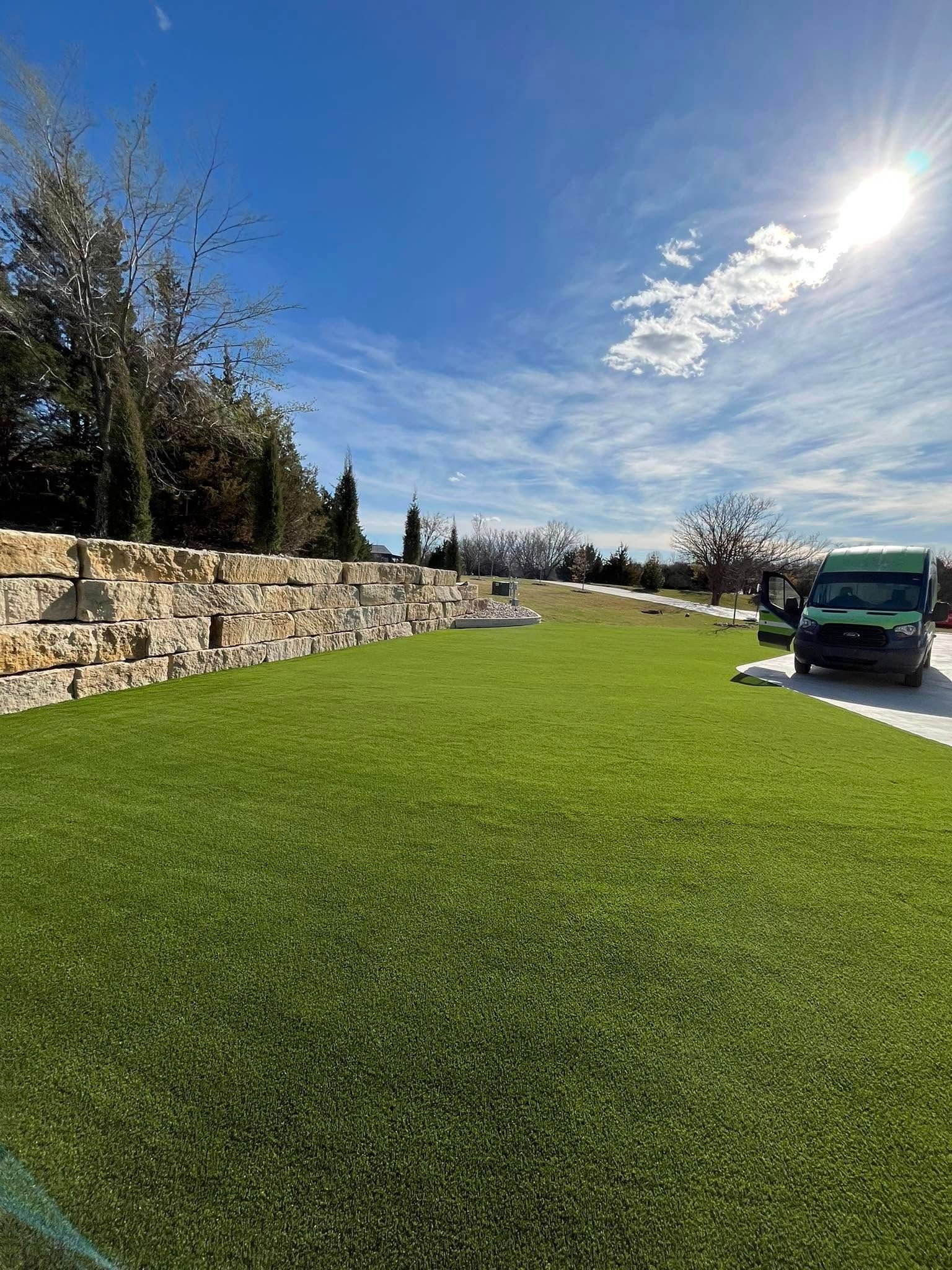 Green lawn next to stone wall under a bright blue sky with a van parked on the right.