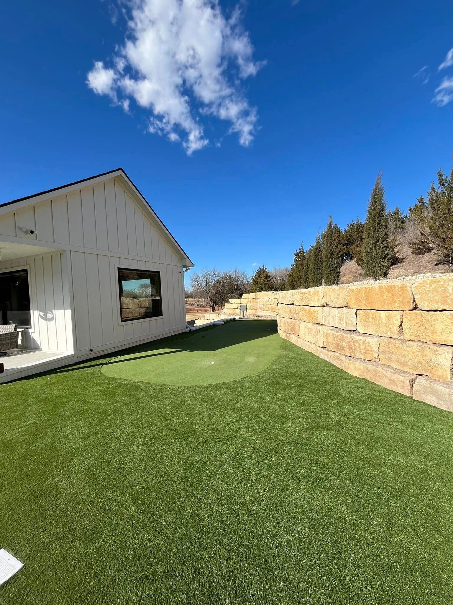 White house with green artificial turf and putting green, blue sky.
