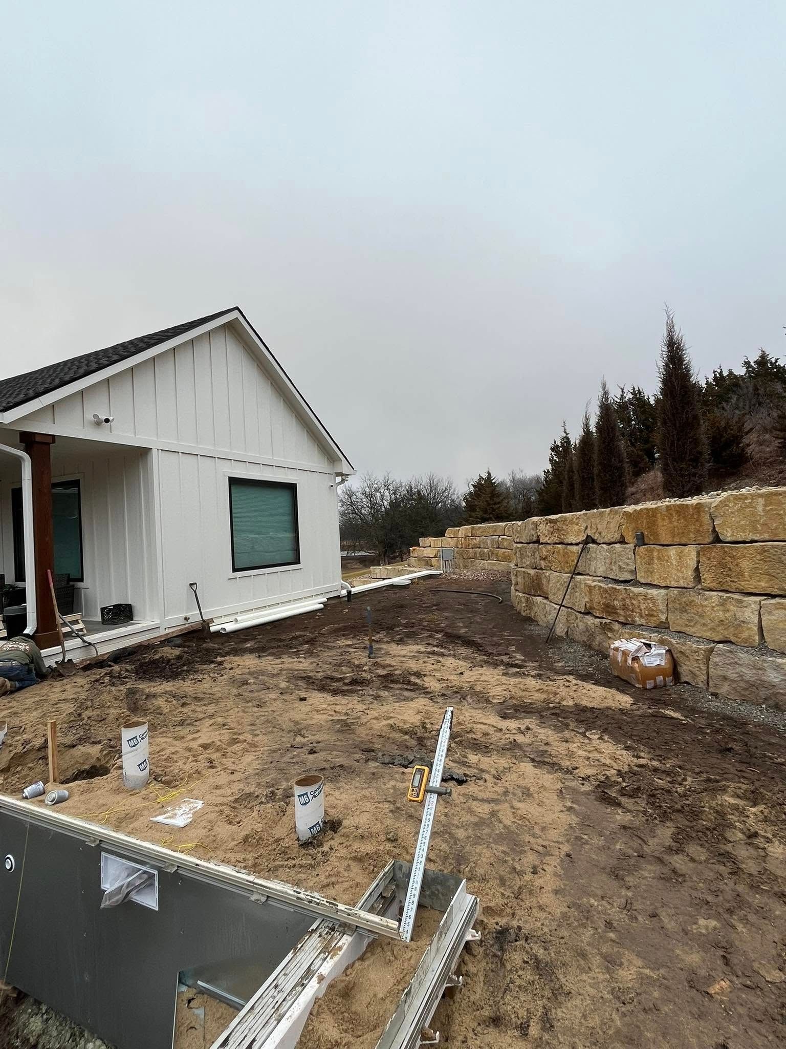 White house under construction with retaining wall, dirt yard, and cloudy sky.