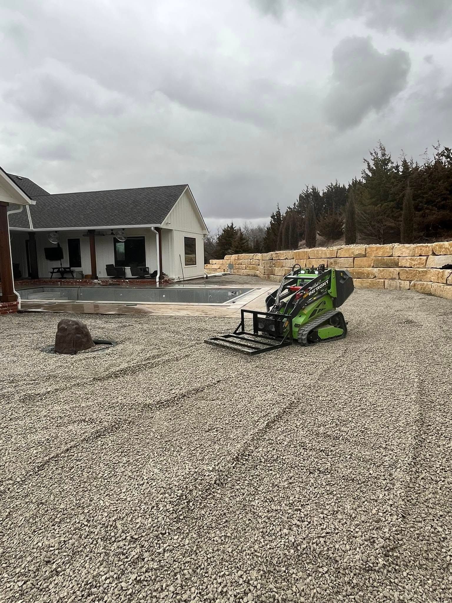 Green skid steer leveling gravel near a home with a pool under a cloudy sky.