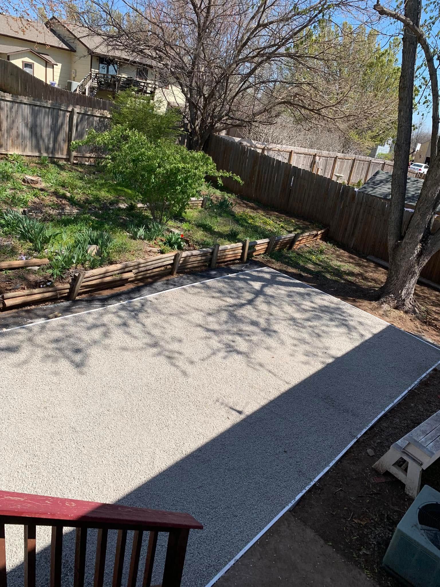 Gravel patio area in a backyard, with a wooden fence and greenery.