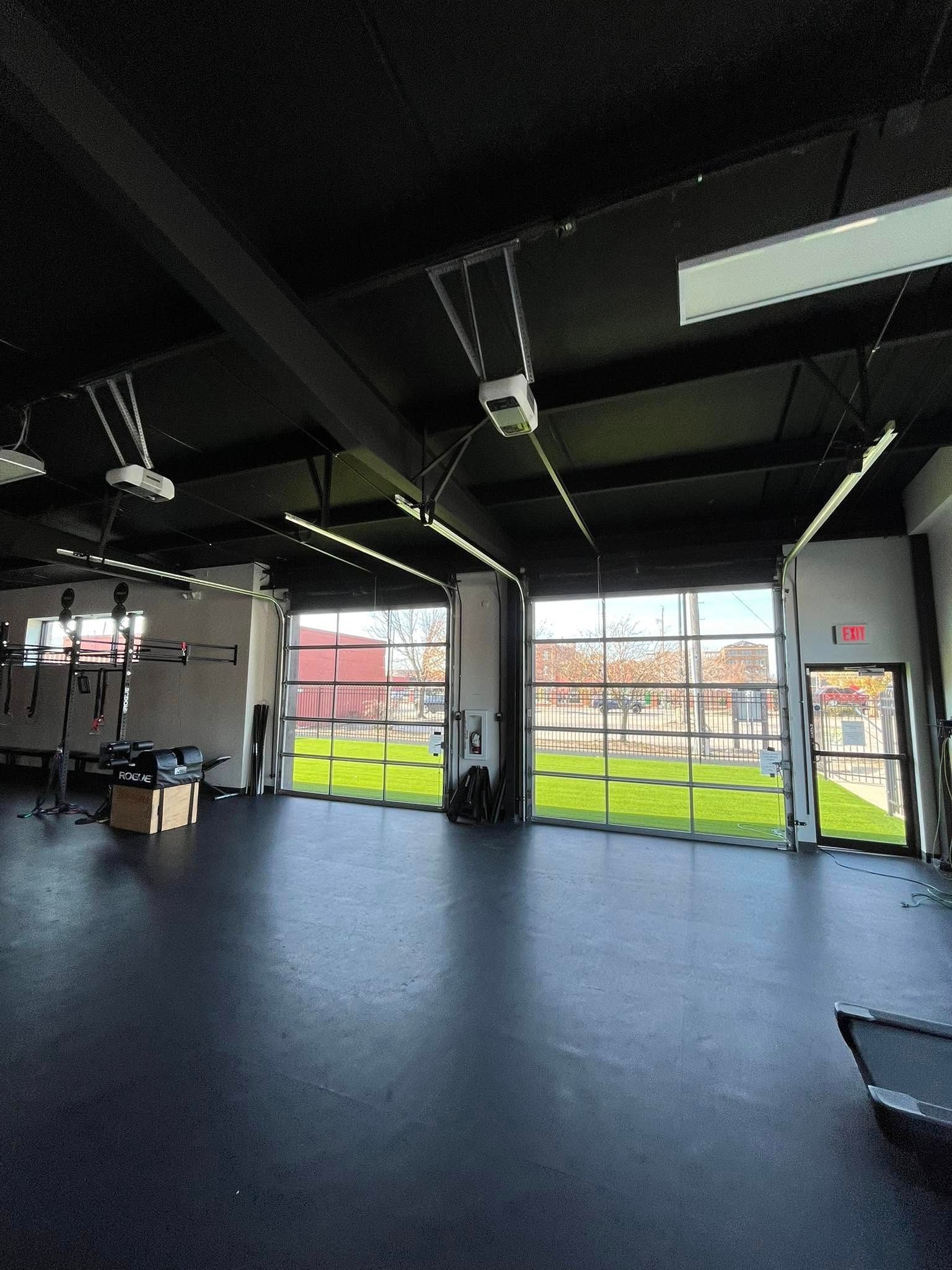 Interior gym space with open glass doors to a grassy area; black ceiling, dark flooring, fitness equipment.