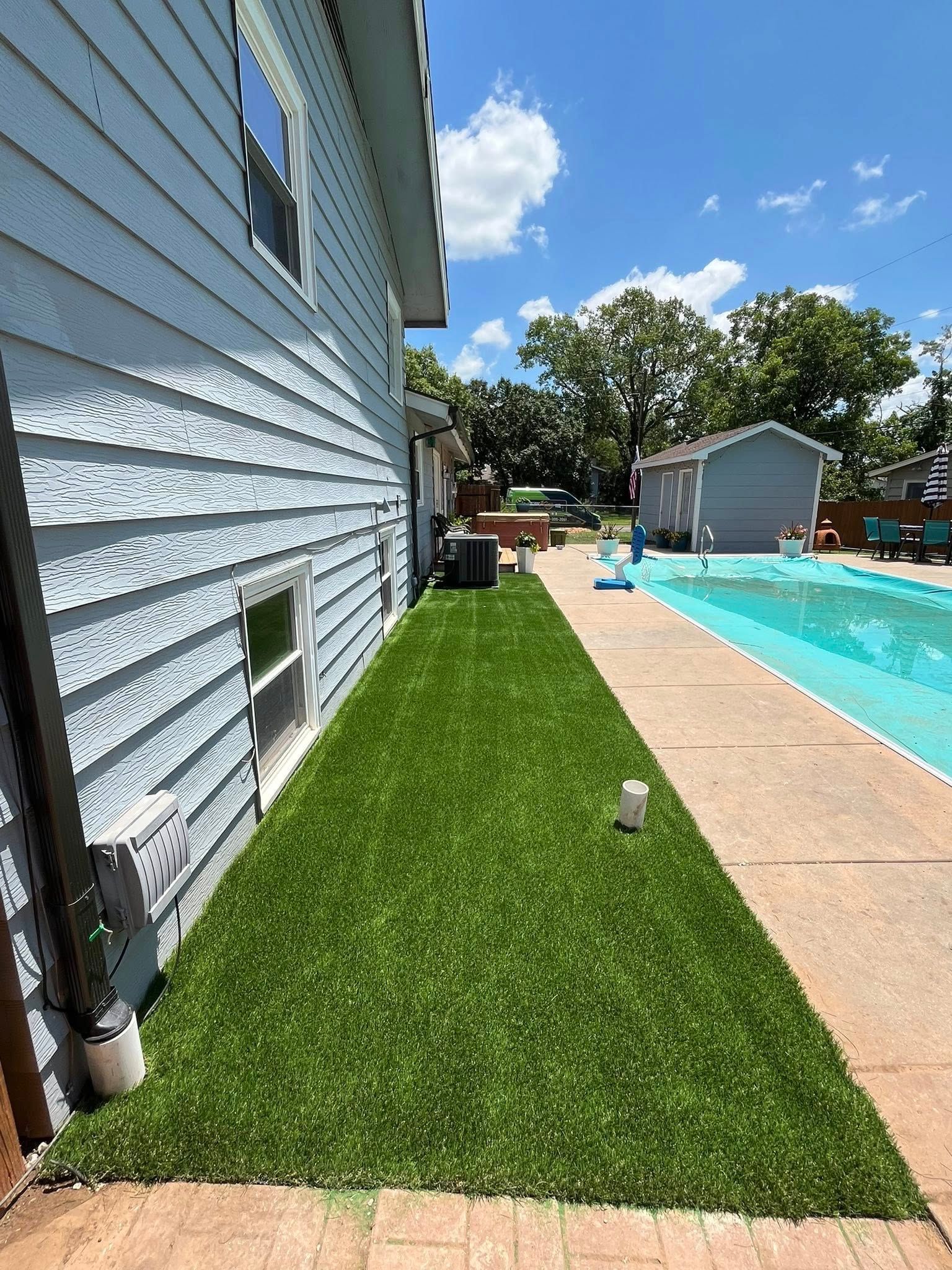 Side view of a house with blue siding and a strip of green artificial turf next to a swimming pool on a sunny day.