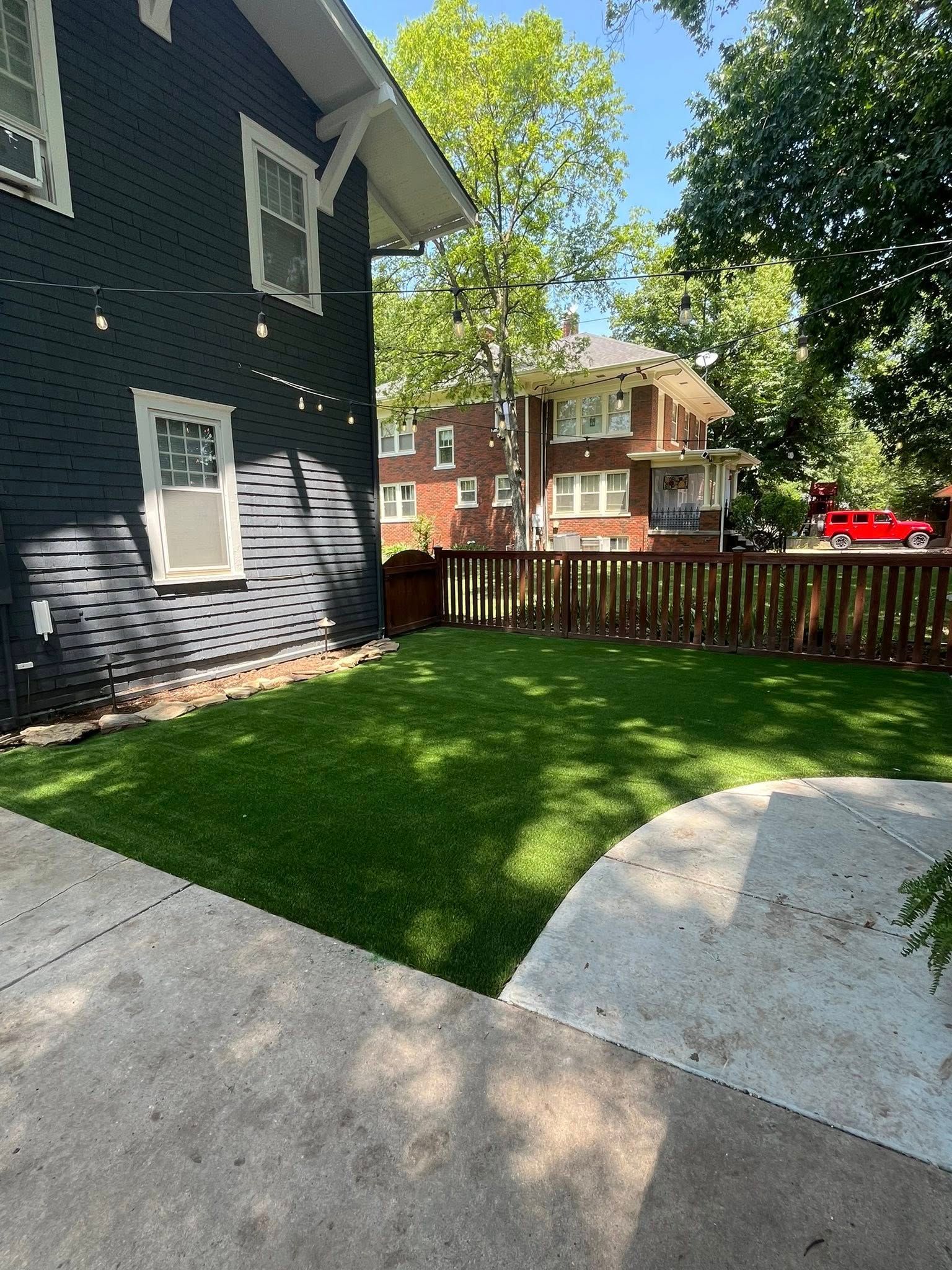 Side view of a dark-painted house, a green lawn, a wooden fence, and other houses in the background.