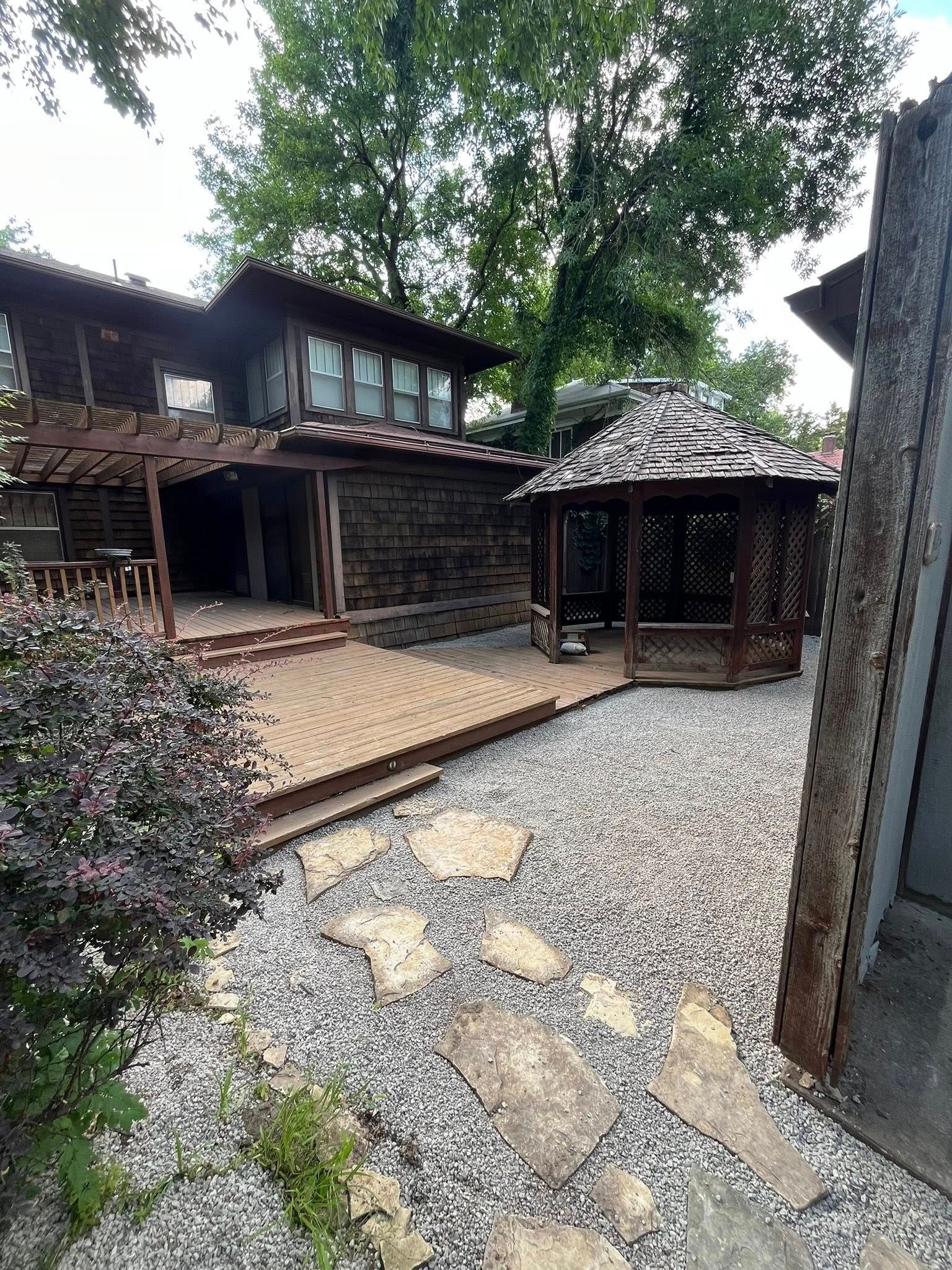 Backyard with a wooden house, gazebo, stone path, and gravel. Brown and green colors.