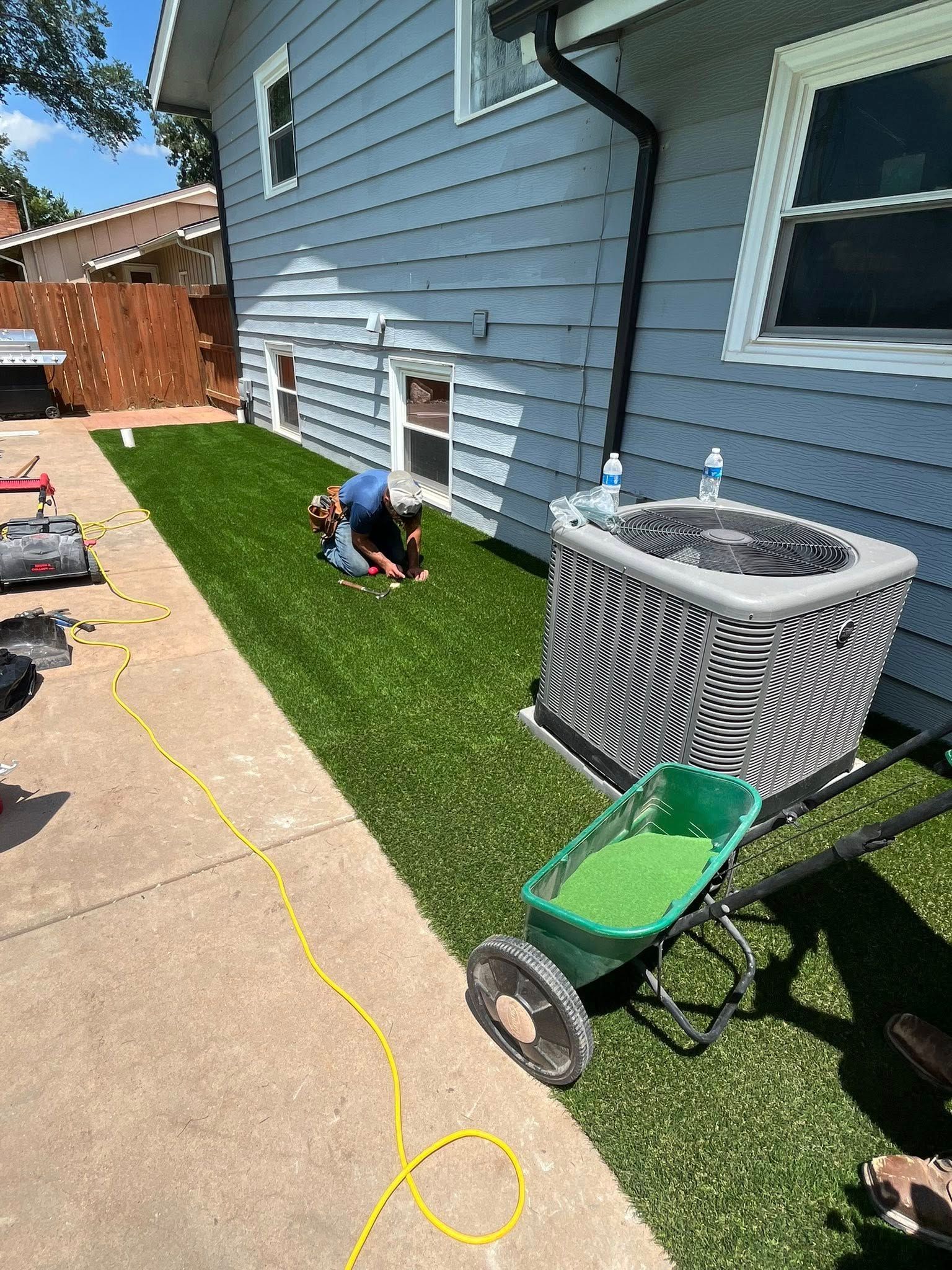 Person installing artificial turf next to a house, with a spreader cart nearby.