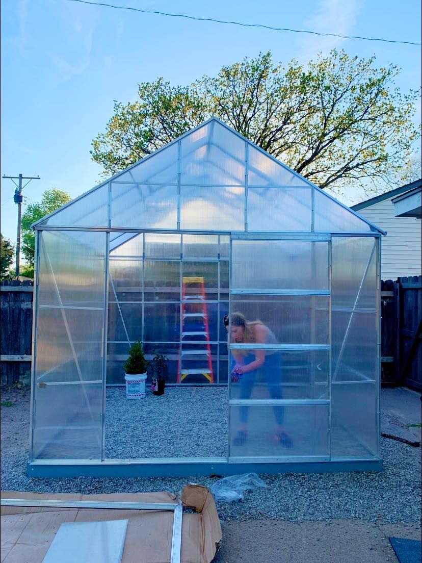 A person inside a partially built greenhouse with a ladder, surrounded by gravel and the sky.