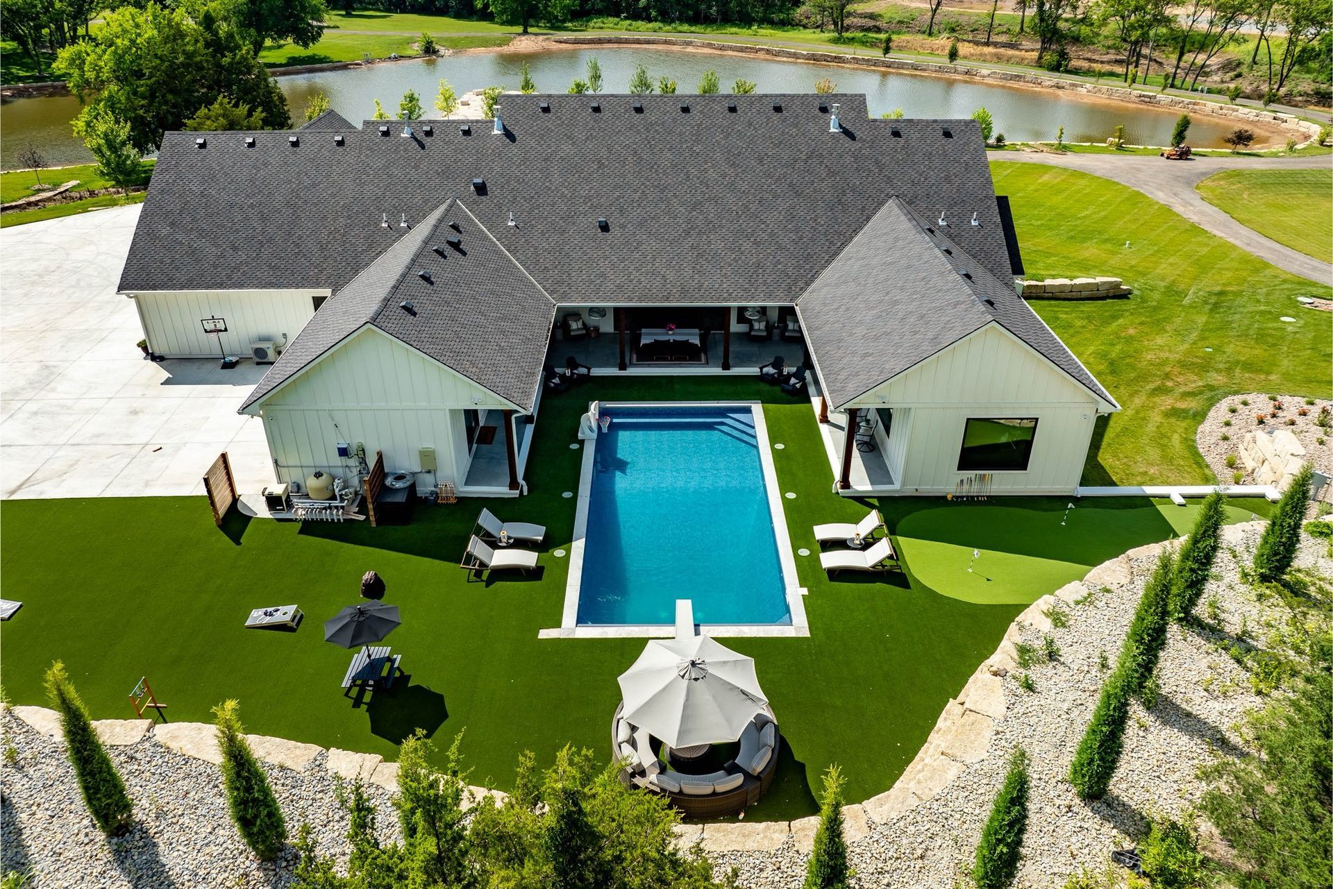 Aerial view of a modern white house with a pool, surrounded by green grass, a pond, and trees.