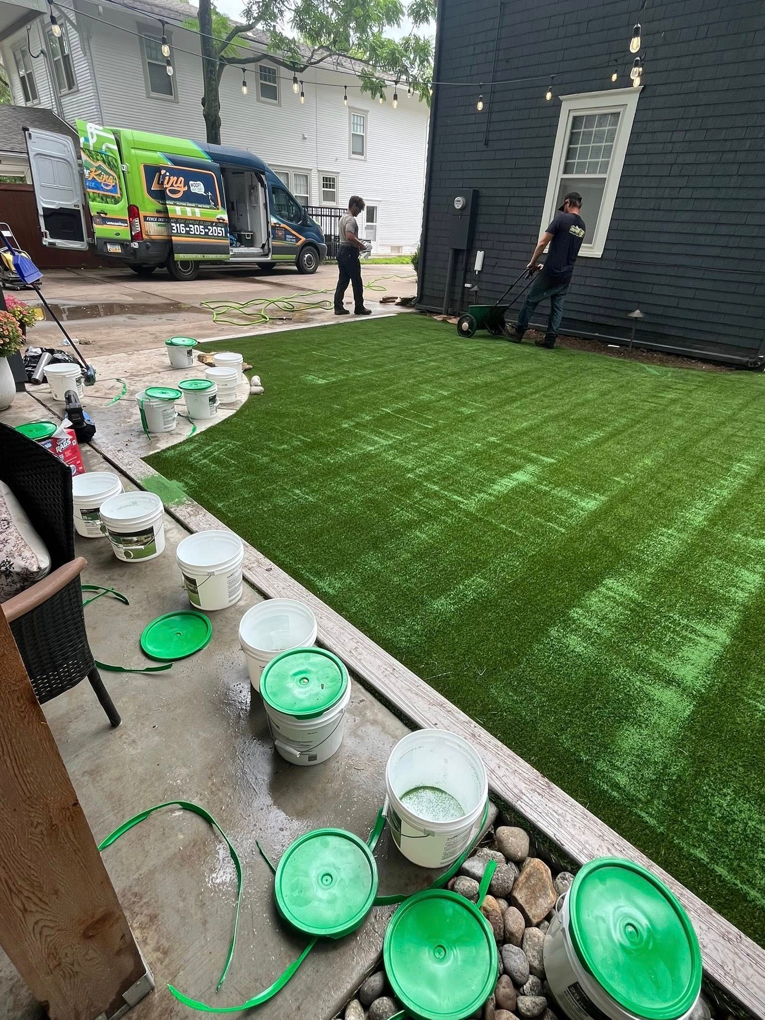 People painting green artificial turf outdoors, surrounded by paint buckets and a van.
