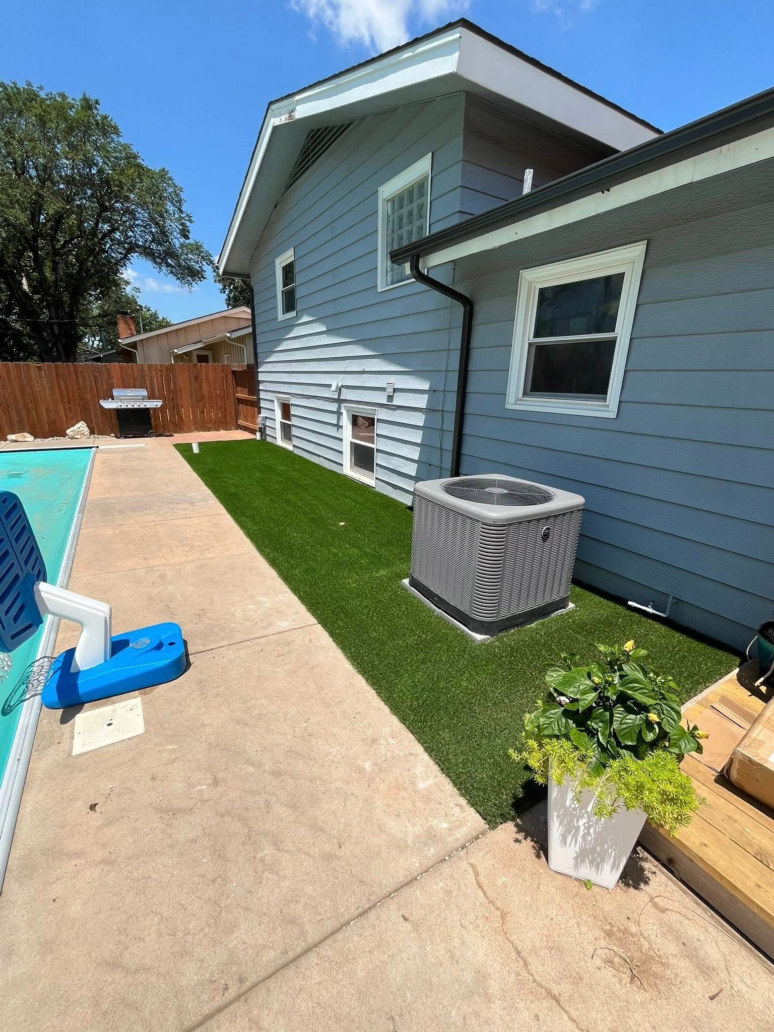 Backyard with pool, blue house, green turf, AC unit, and potted plant.